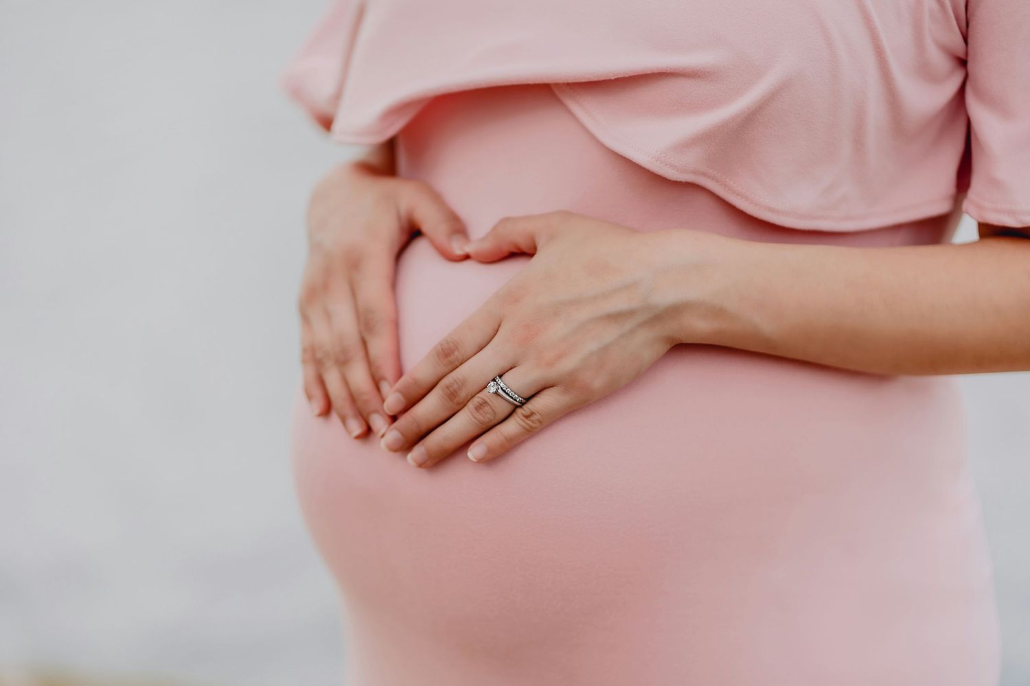 Pregnant woman in pink dress cradling baby bump with both hands, wearing a wedding ring, close-up maternity photo.