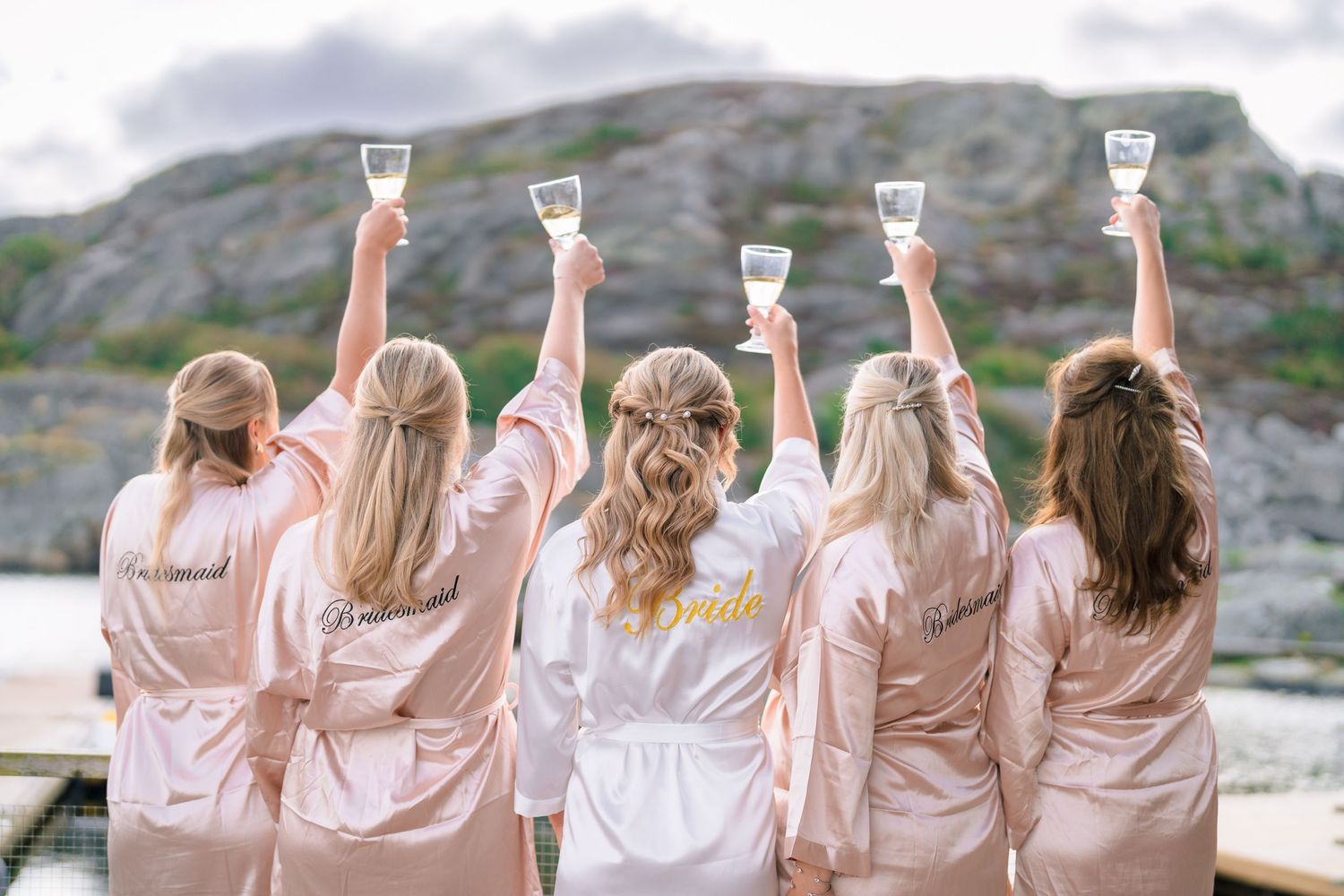 Bridesmaids in matching blush satin robes raising champagne glasses outdoors with rocky coastal scenery behind them.
