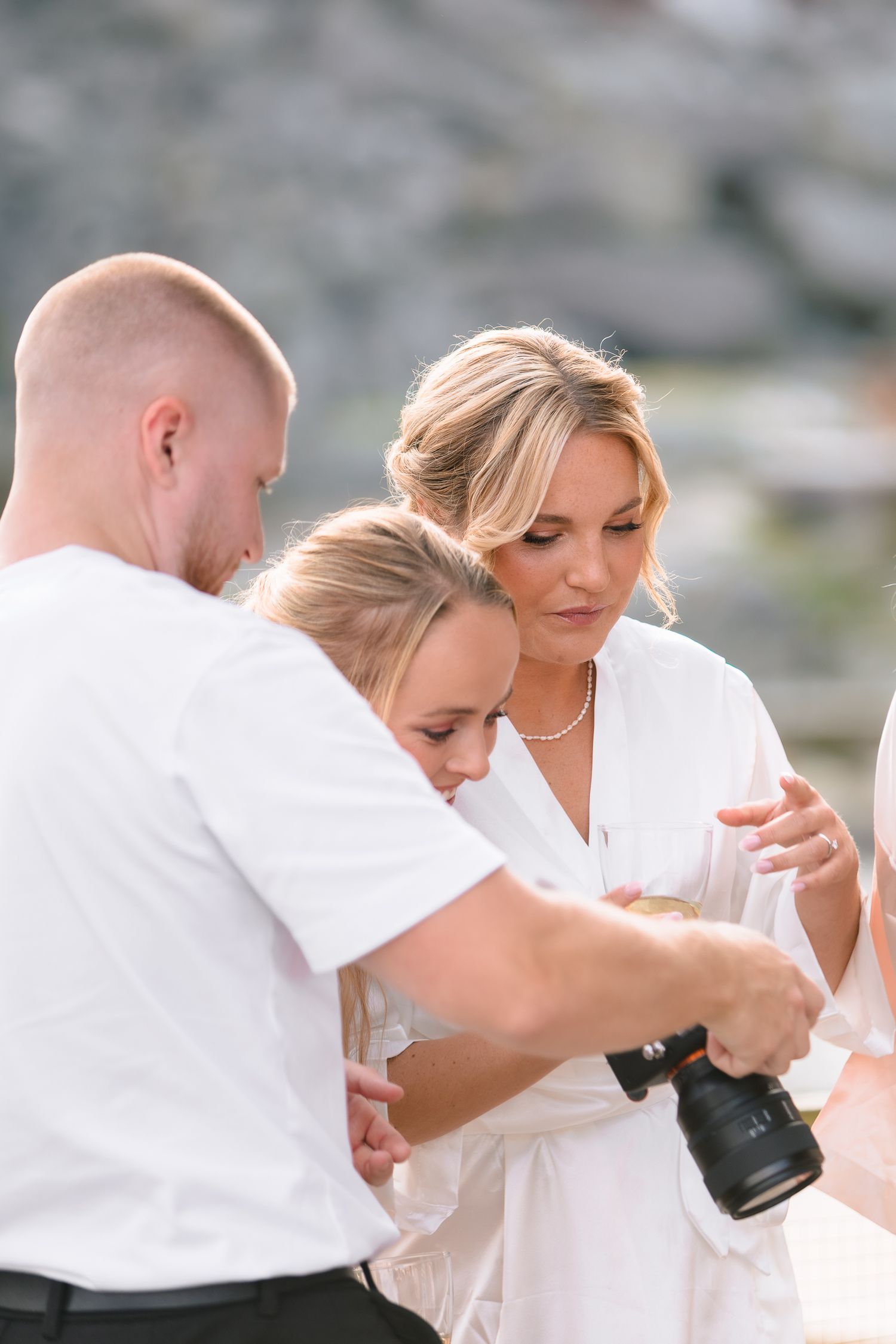 Three people in white attire reviewing photos on a DSLR camera outdoors during a bright wedding shoot.
