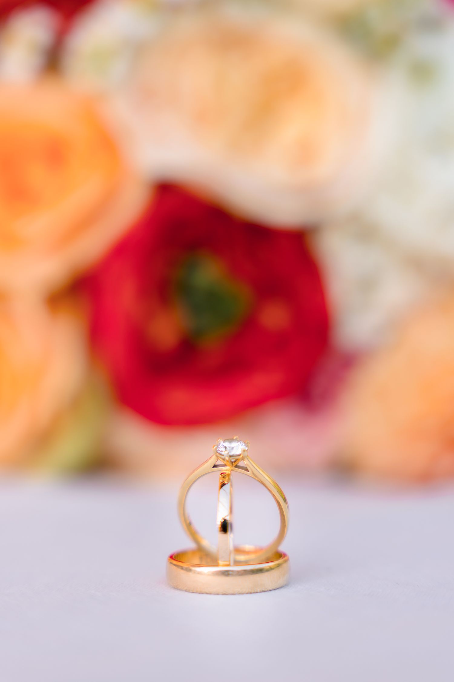 Gold wedding band and diamond solitaire engagement ring standing upright before floral bouquet.