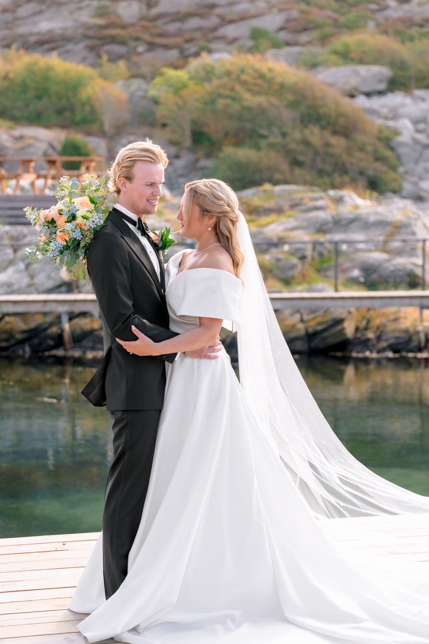Bride and groom embrace on a dock by the water, she in an off-shoulder gown with veil, he in a black tuxedo.