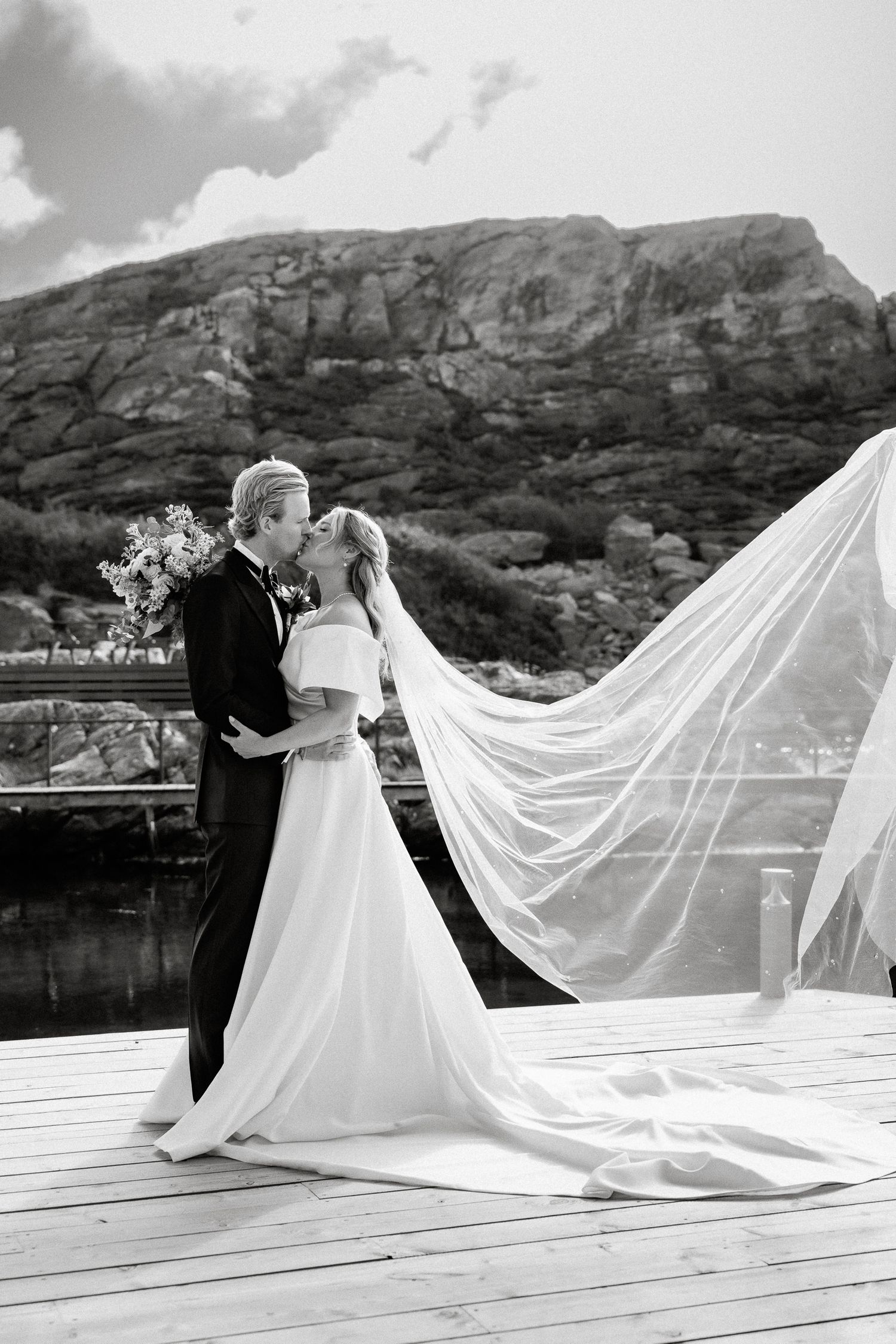 Black and white wedding photo of bride and groom kissing on dock, veil flowing in wind with rocky mountain backdrop.