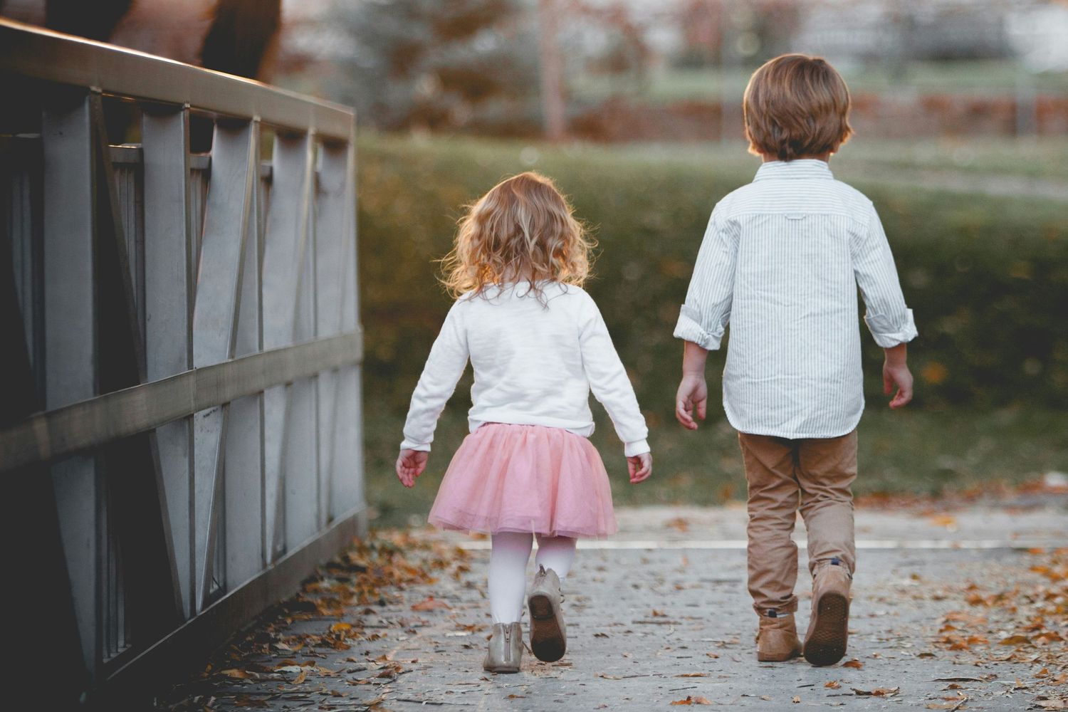 Two young children walking hand in hand on a path beside a bridge, girl in pink tutu, boy in tan pants.