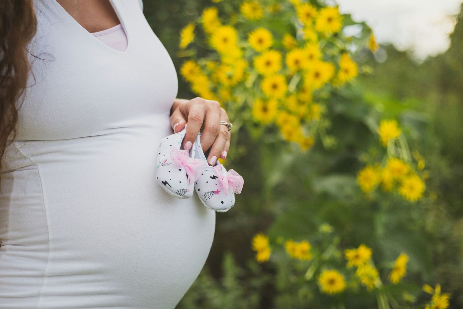 Pregnant woman in white dress holding tiny pink baby booties with yellow sunflowers in the background.