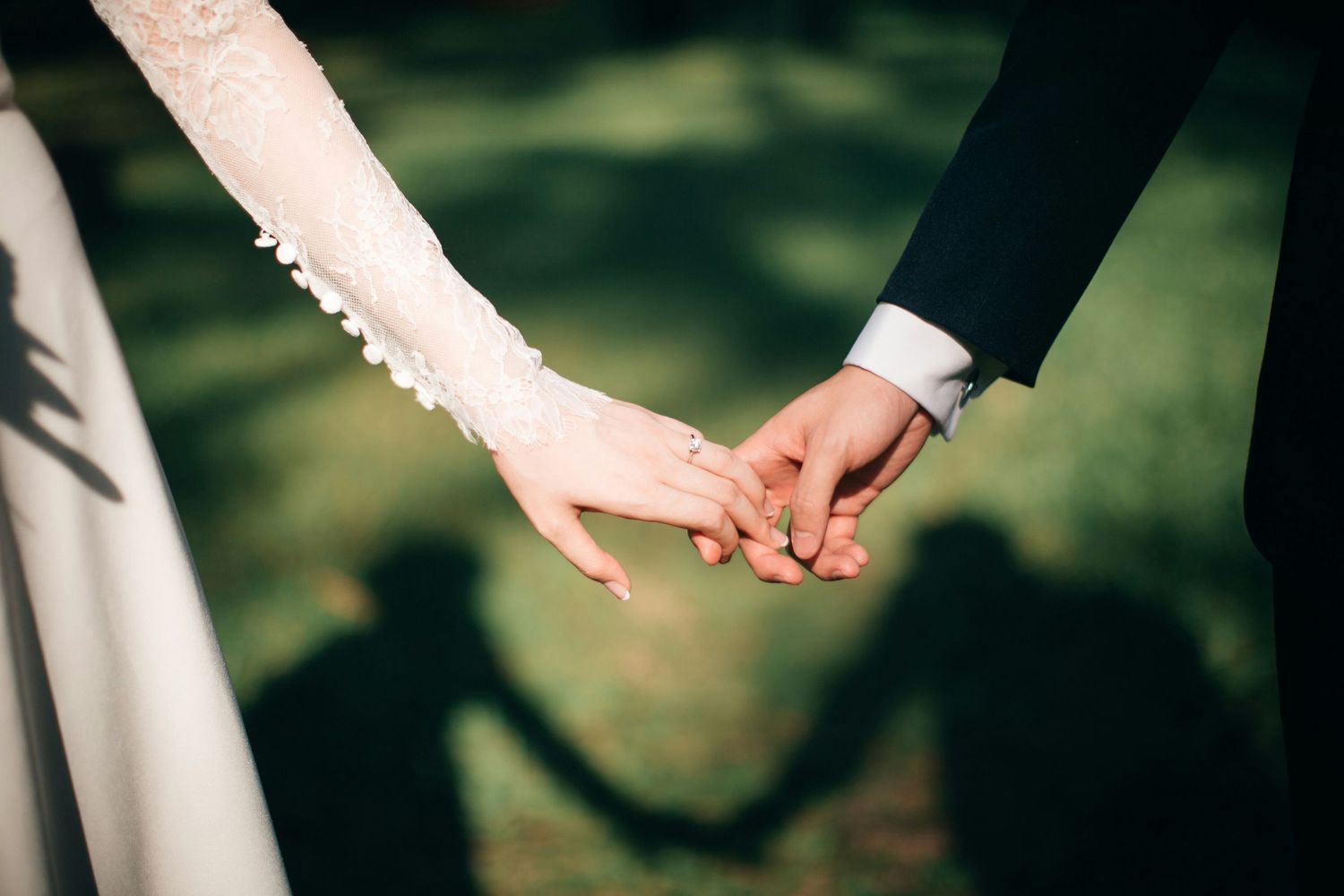 Bride and groom holding hands at wedding ceremony, casting romantic shadows on green grass.