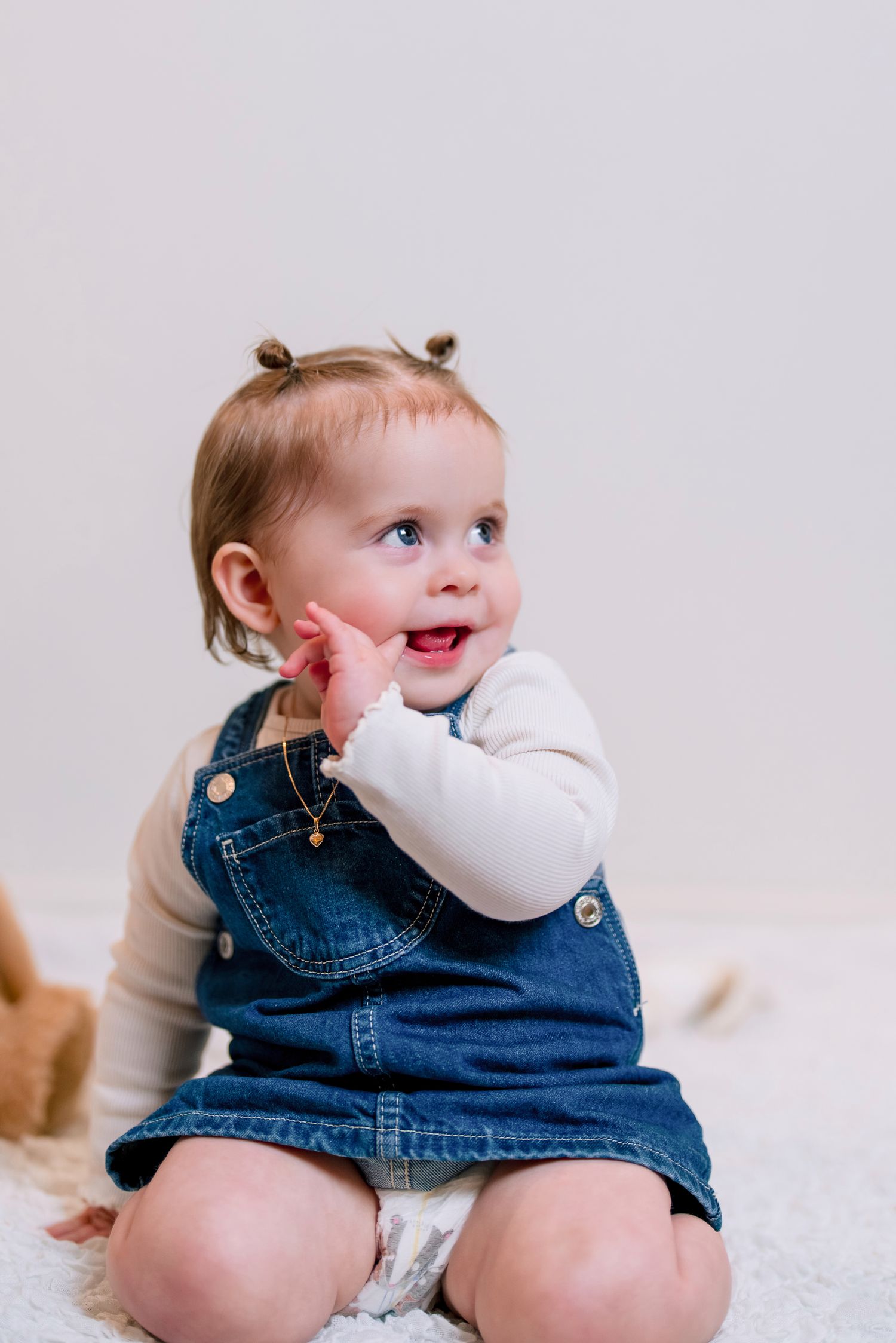 Smiling baby girl in denim overalls and white shirt sitting on a fluffy white rug with hand raised to face.