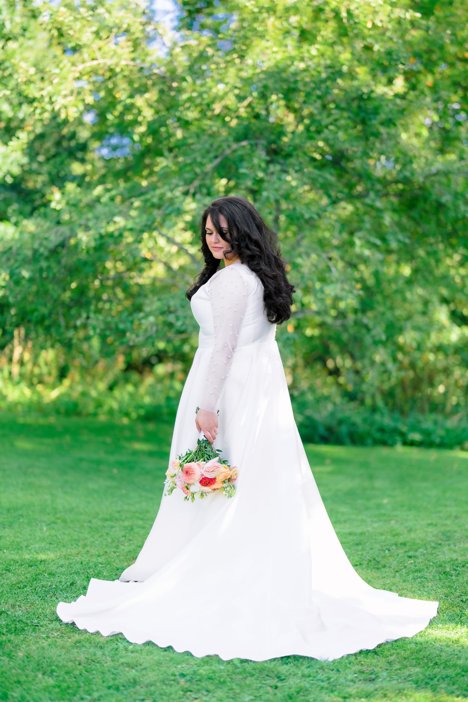 Bride in elegant long-sleeve white lace wedding gown holding floral bouquet standing in lush green garden.