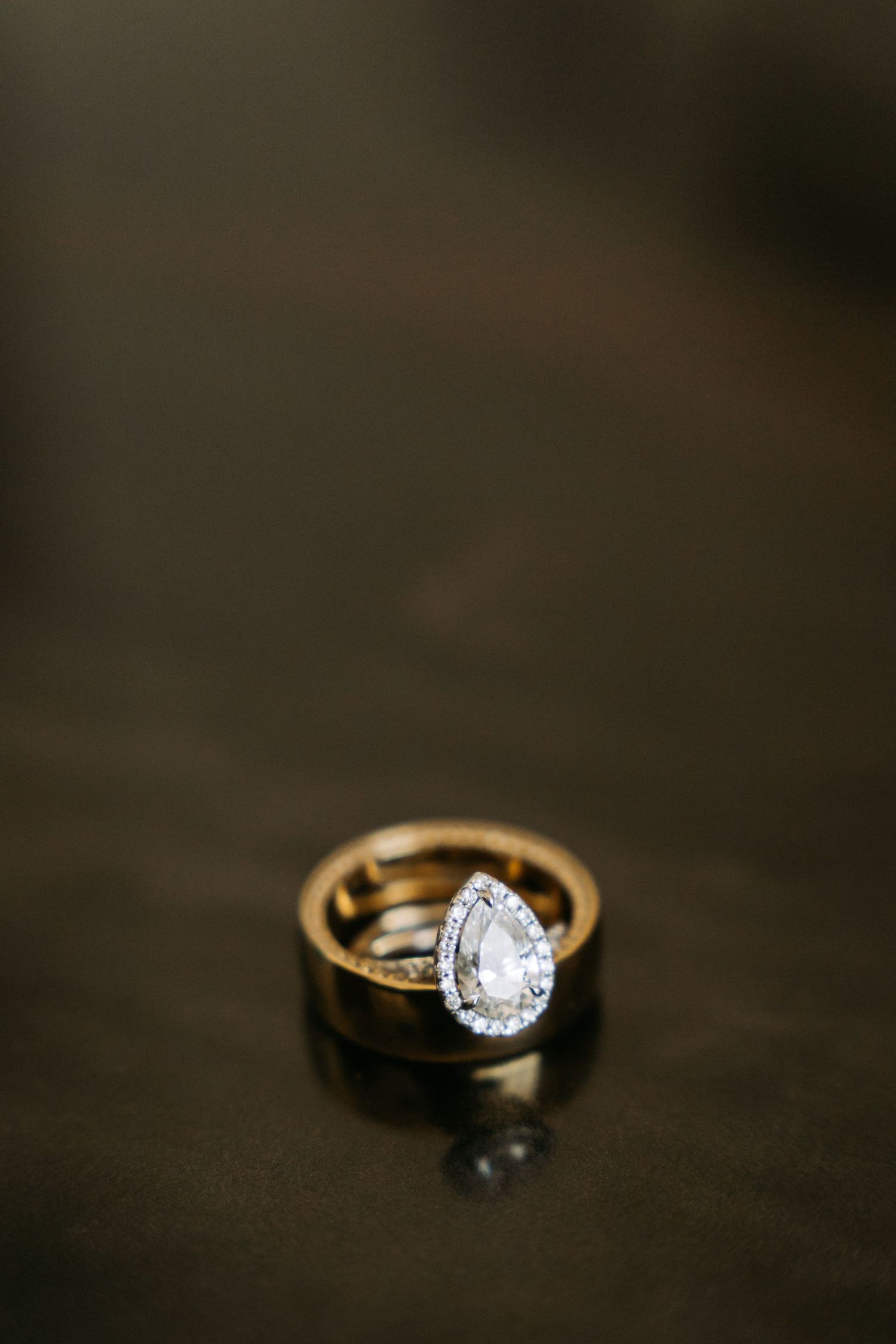 Elegant pear-shaped diamond halo engagement ring resting on gold wedding band on dark reflective surface.