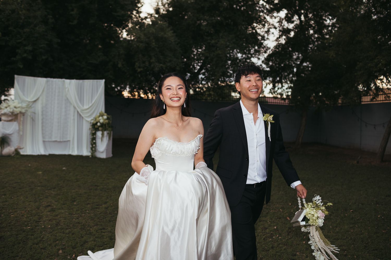 Bride in elegant white ballgown and groom walking outdoors near wedding ceremony backdrop.