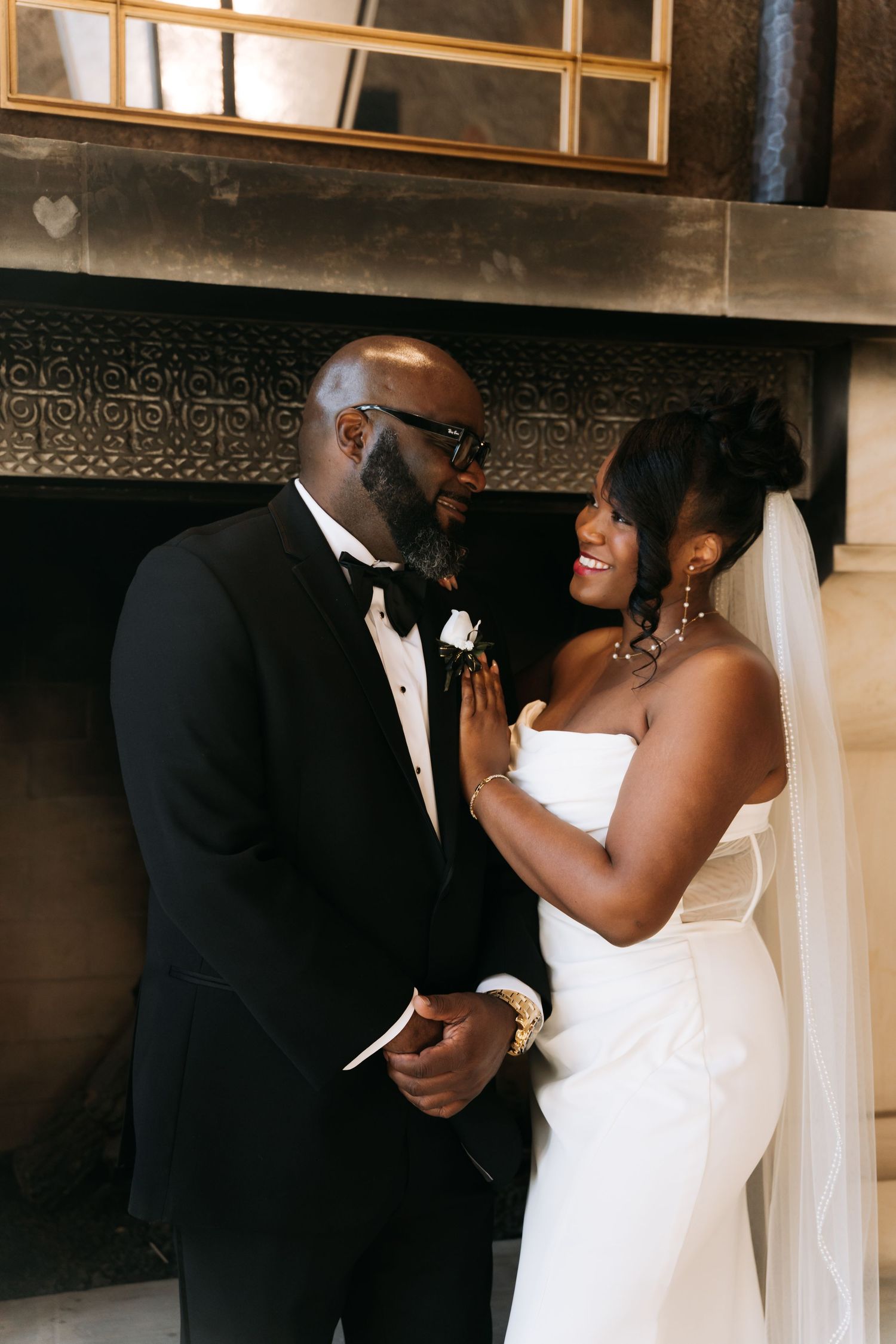 Bride in white strapless gown and groom in black tuxedo sharing a tender moment by fireplace.
