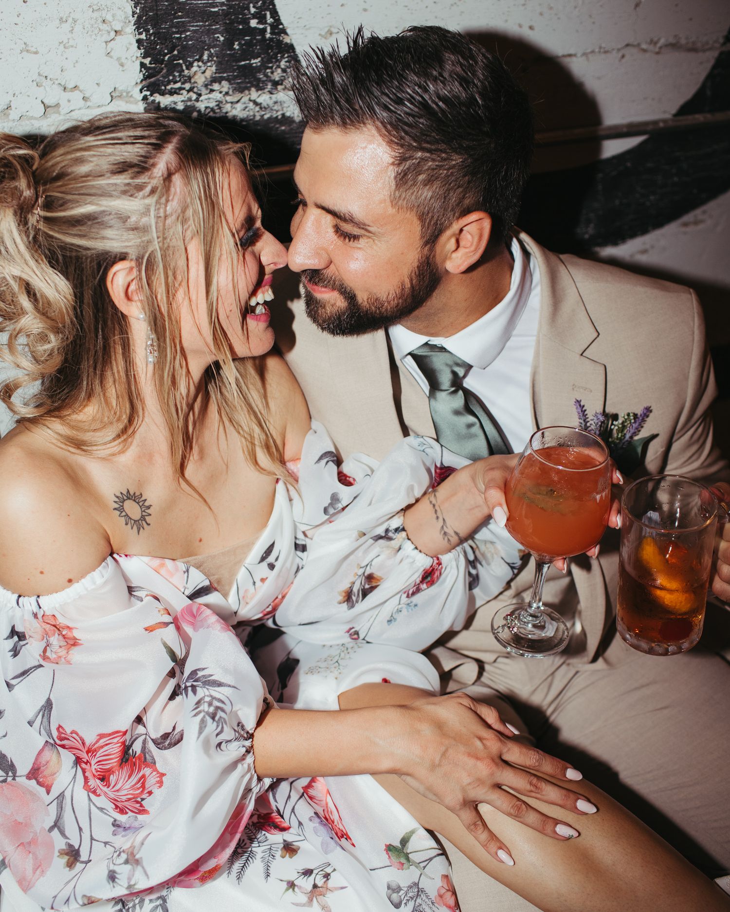 Bride in off-shoulder floral dress and groom in tan suit sharing an intimate moment with drinks.