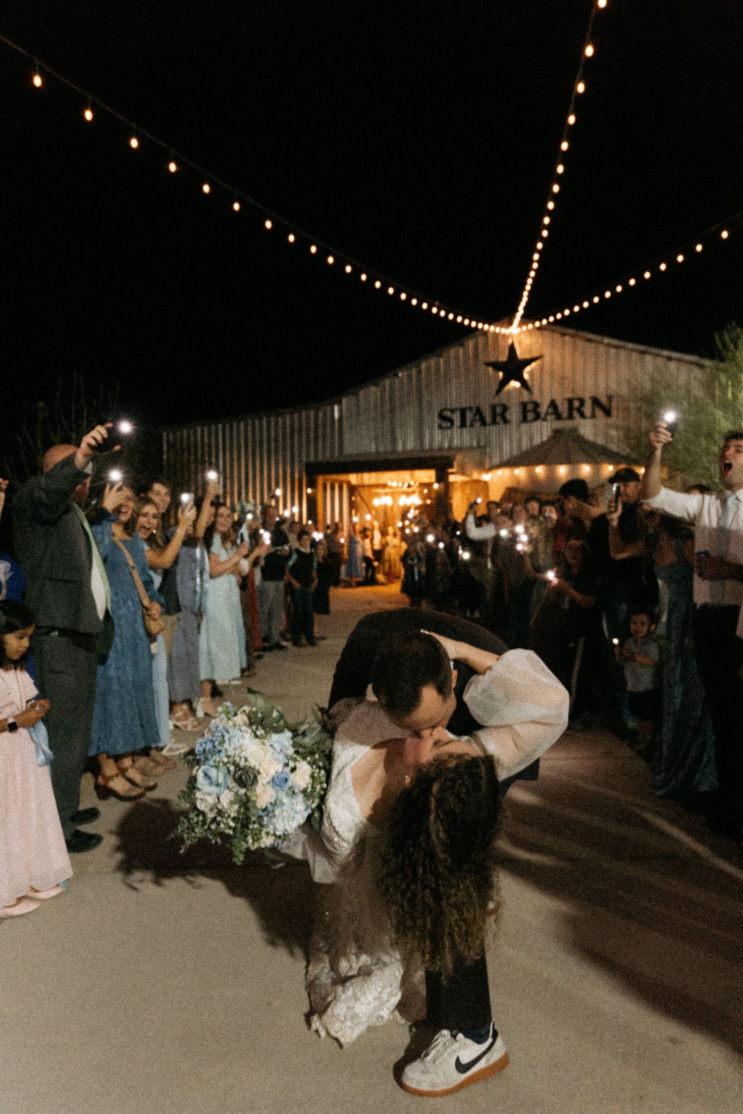 Groom dips bride for kiss during nighttime wedding exit at Star Barn venue with flashlights and string lights.