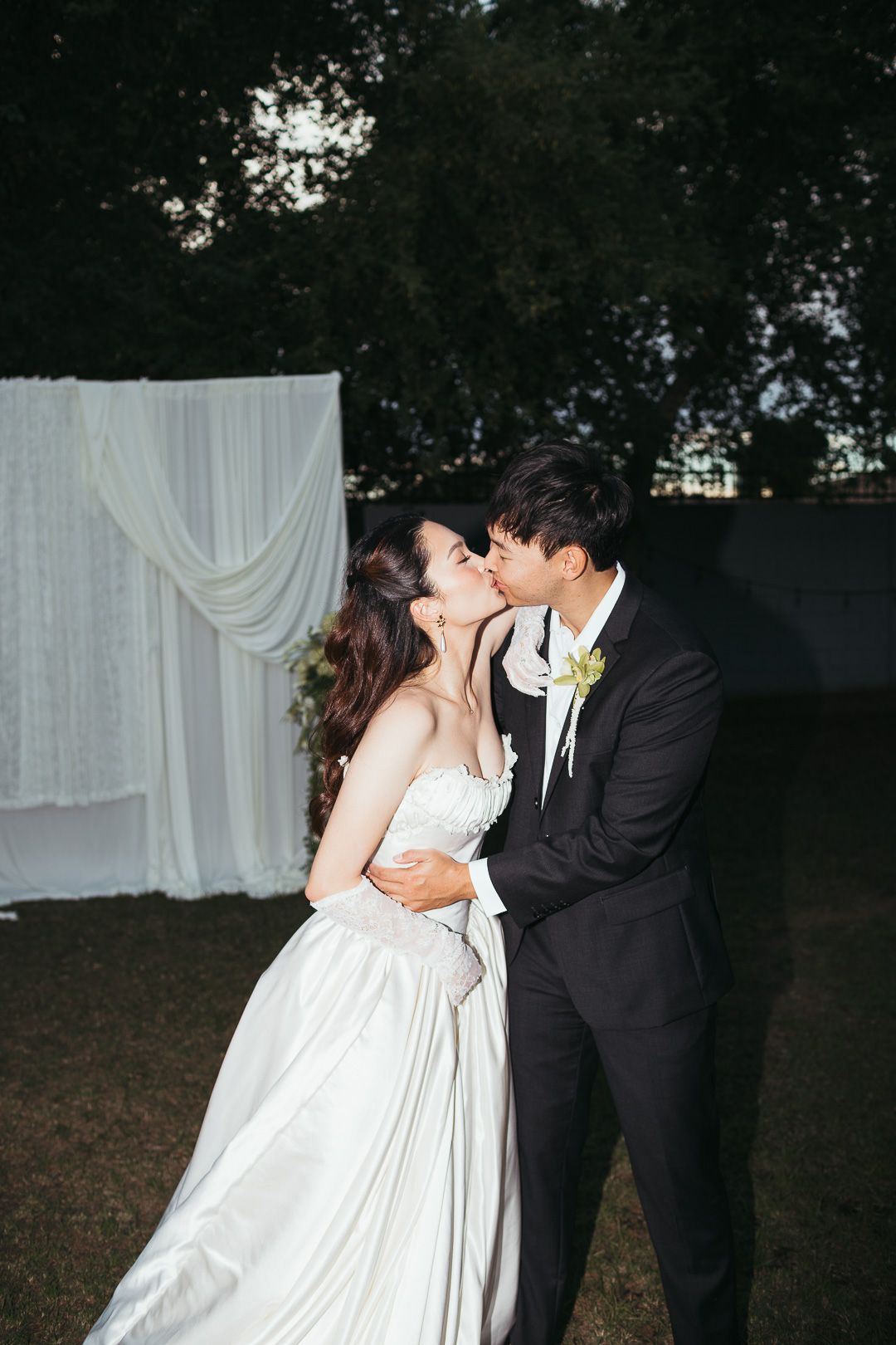 Bride and groom share a romantic kiss outdoors in front of a white draped wedding backdrop at dusk.