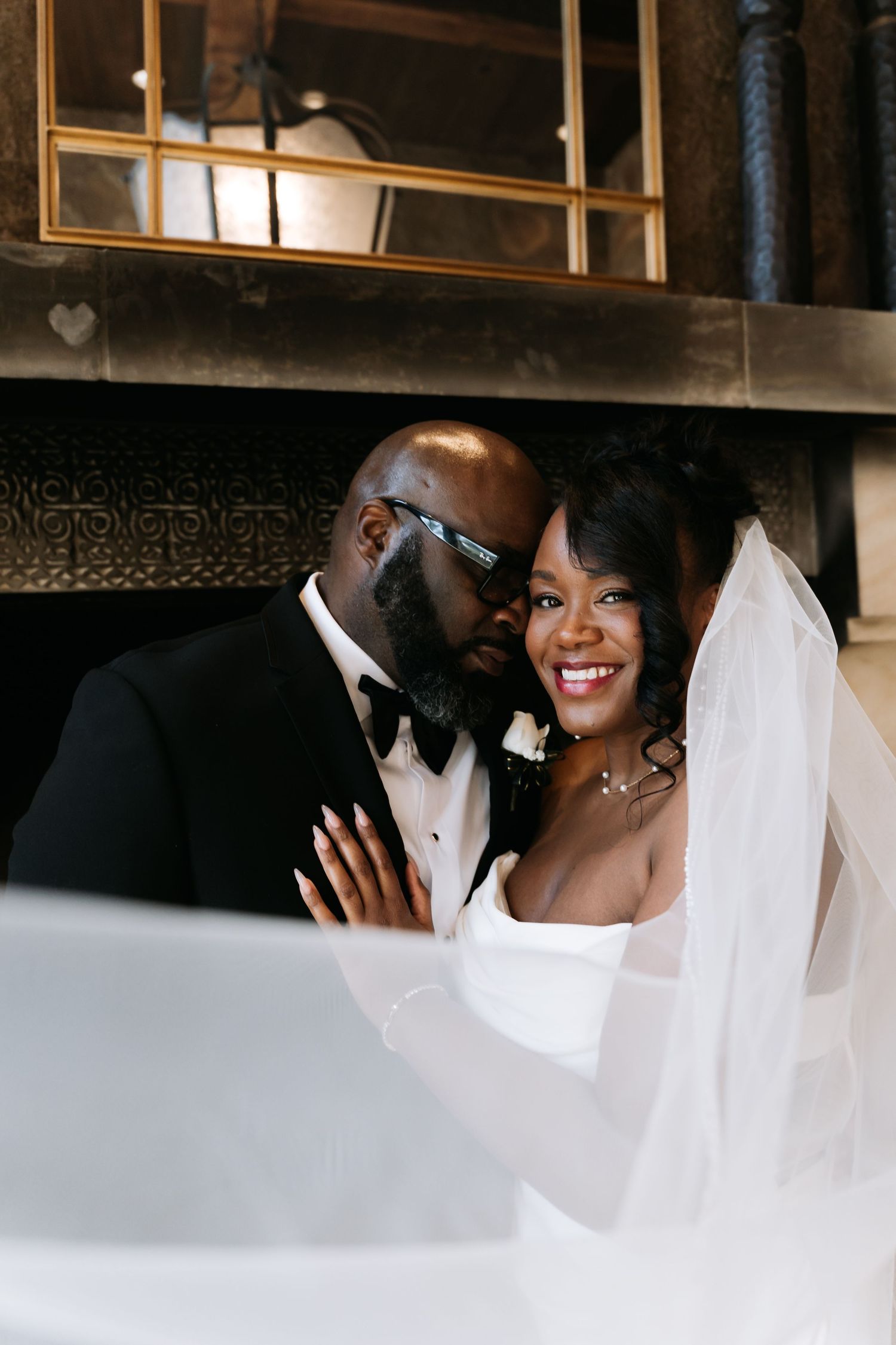 Groom in tuxedo whispers to smiling bride in white gown and veil in warmly lit wedding venue.