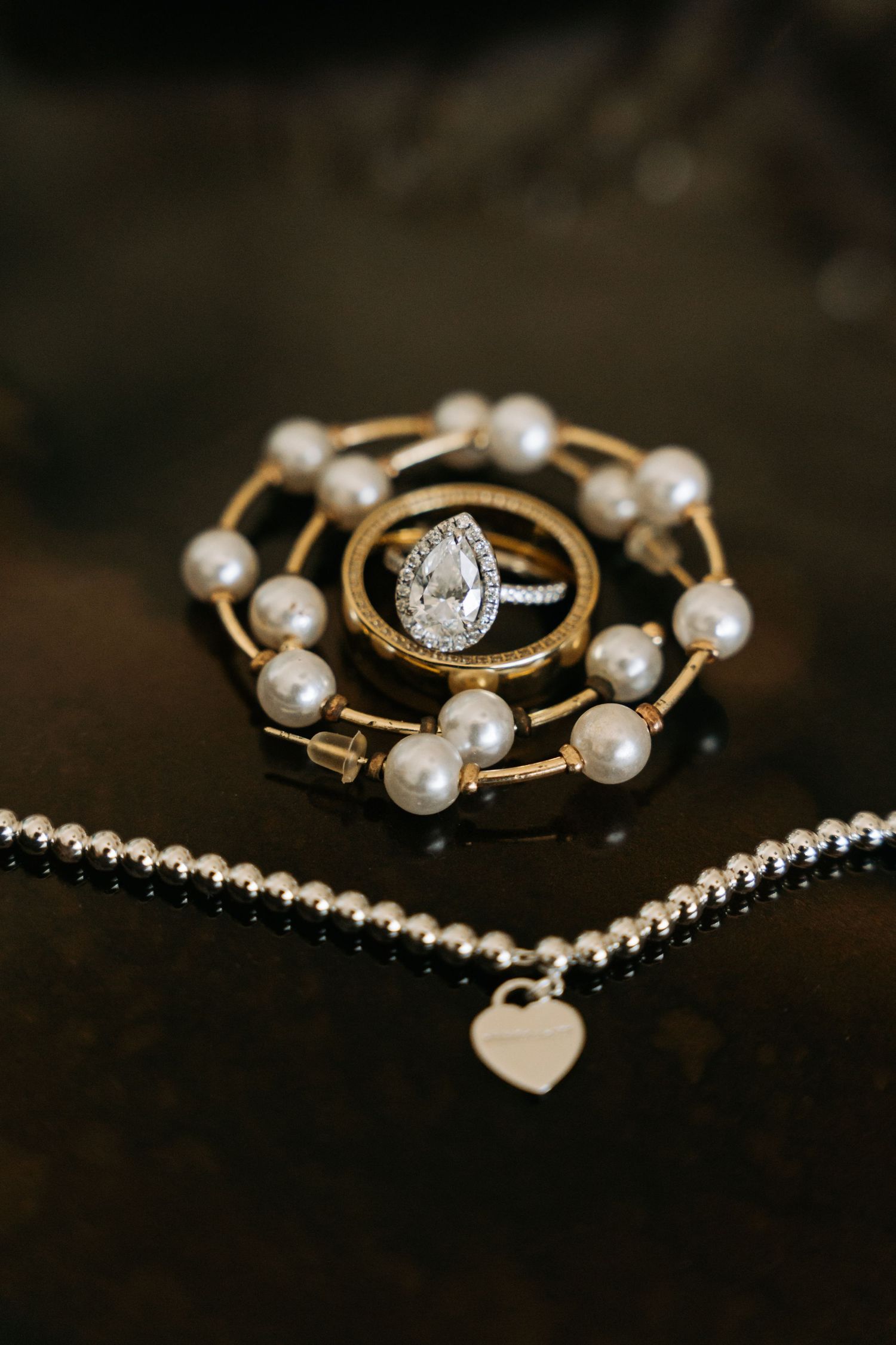 Pear diamond engagement ring displayed on pearl bracelet beside silver heart charm necklace on dark surface.