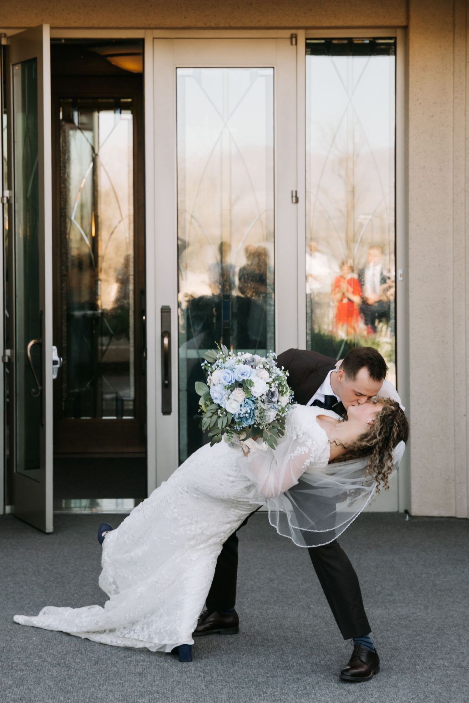 Groom dips bride in a romantic kiss outside glass doors while she holds a blue and white bouquet.