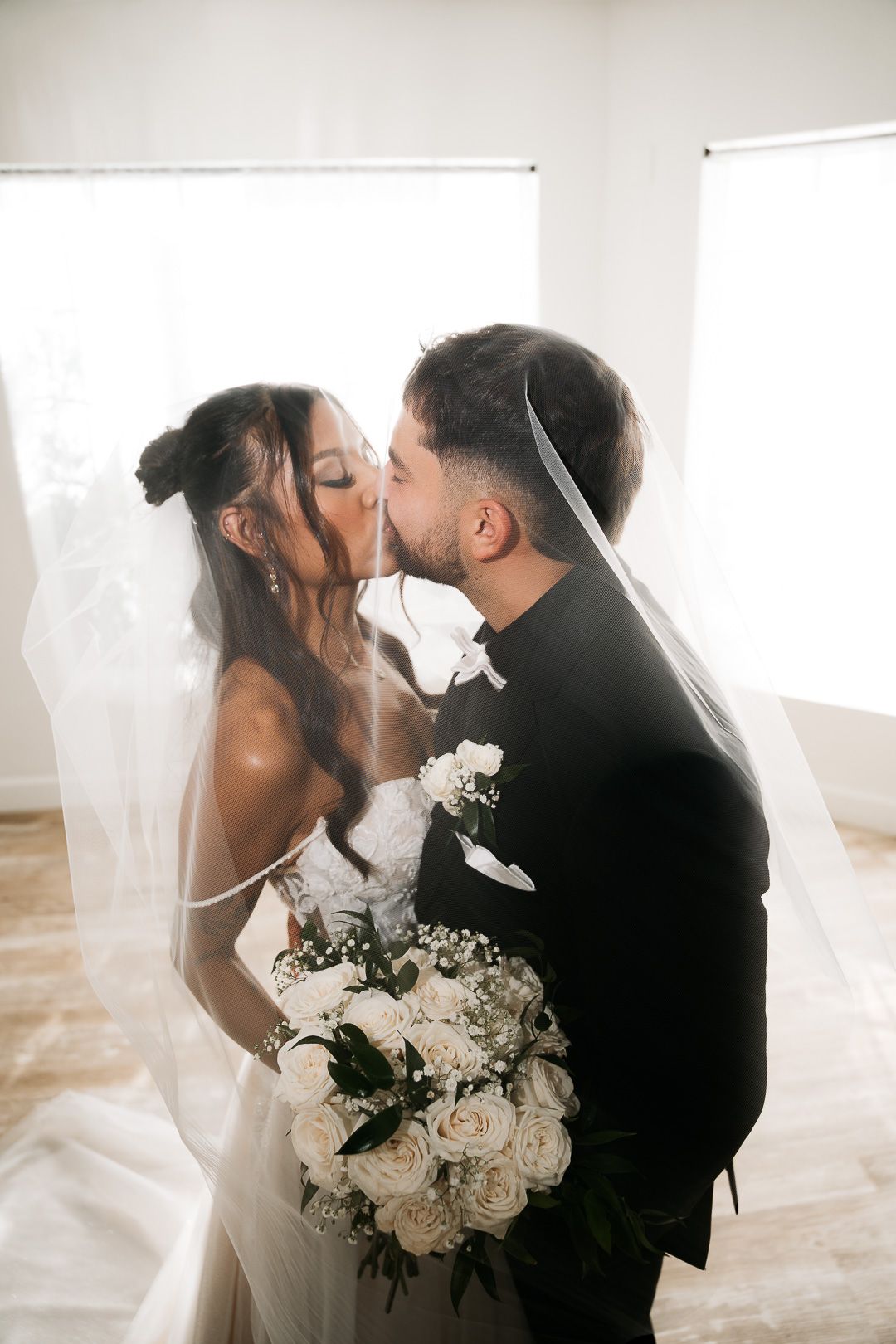 Bride and groom sharing a romantic kiss under a flowing veil while holding a white rose bouquet.