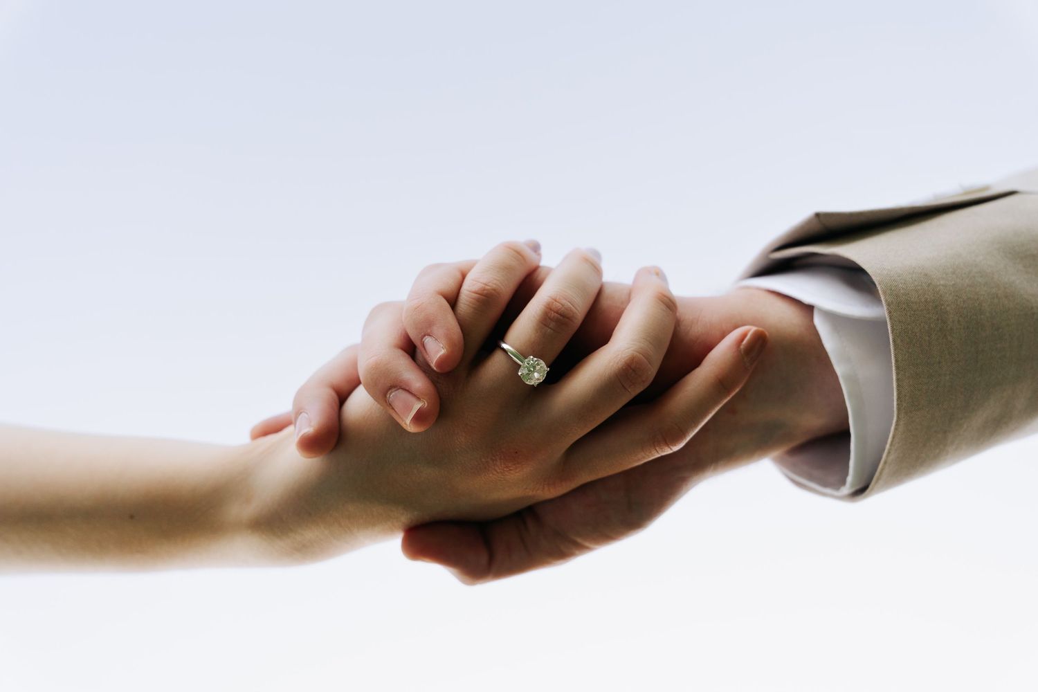 Close-up of couple holding hands showing diamond engagement ring, groom wearing beige suit jacket.