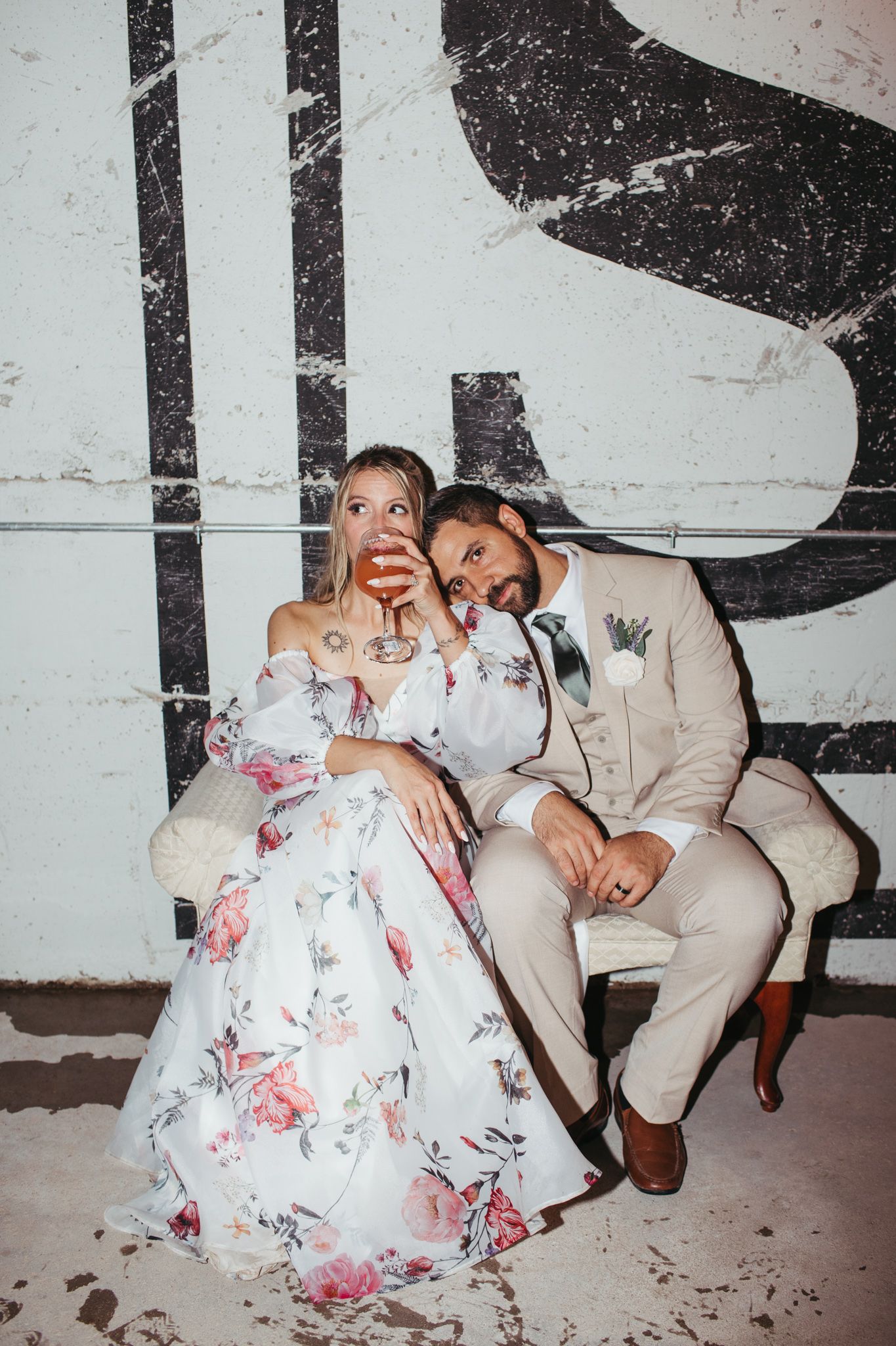 Bride in floral gown and groom in tan suit sitting together in front of a vintage wall mural.