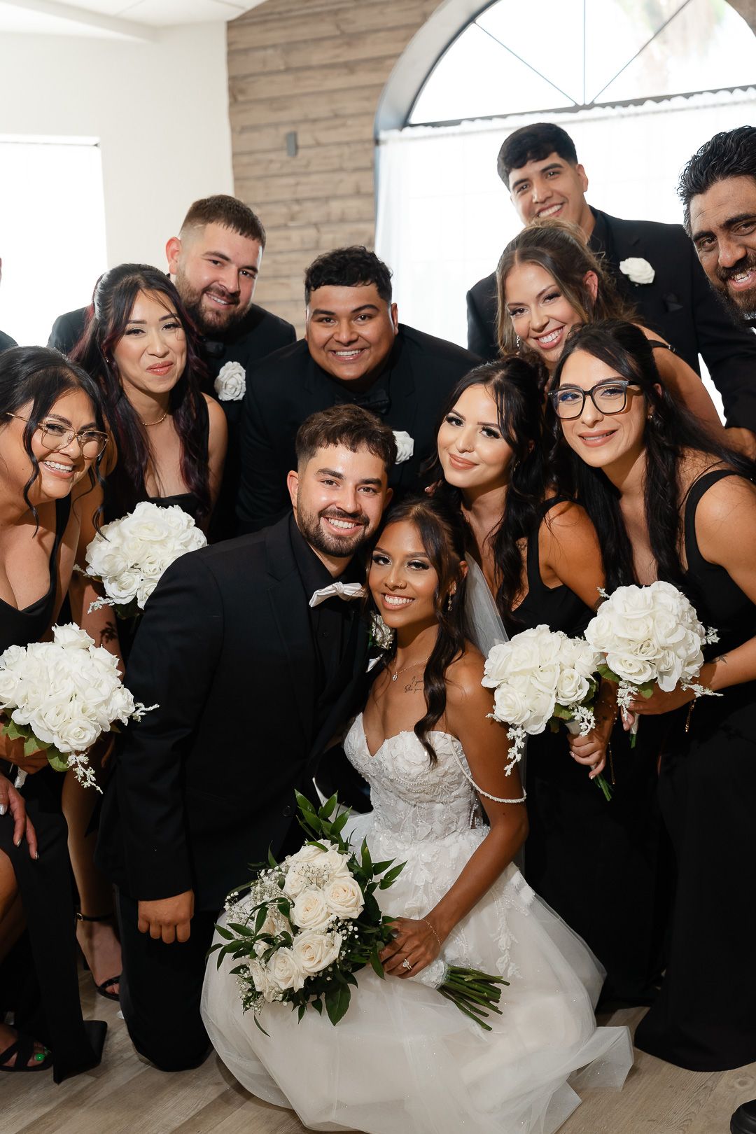 Bride and groom pose with wedding party in matching black attire holding white bouquets indoors.