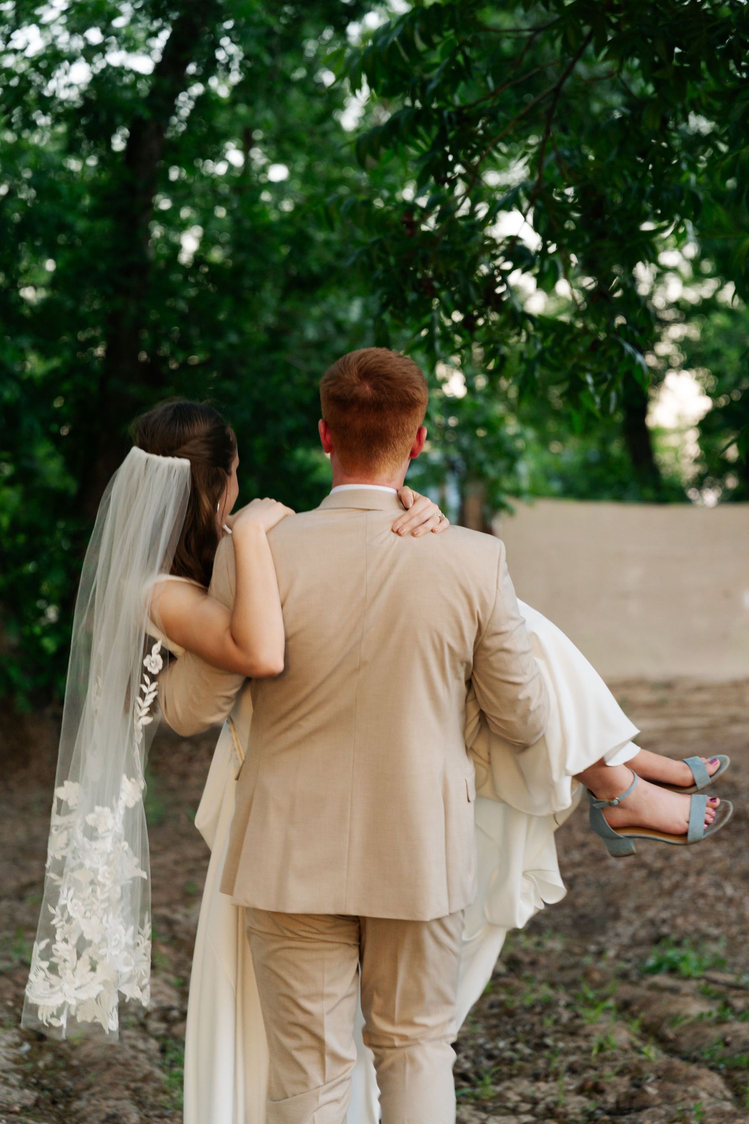 Groom carries bride through green garden setting, both dressed in wedding attire, viewed from behind.