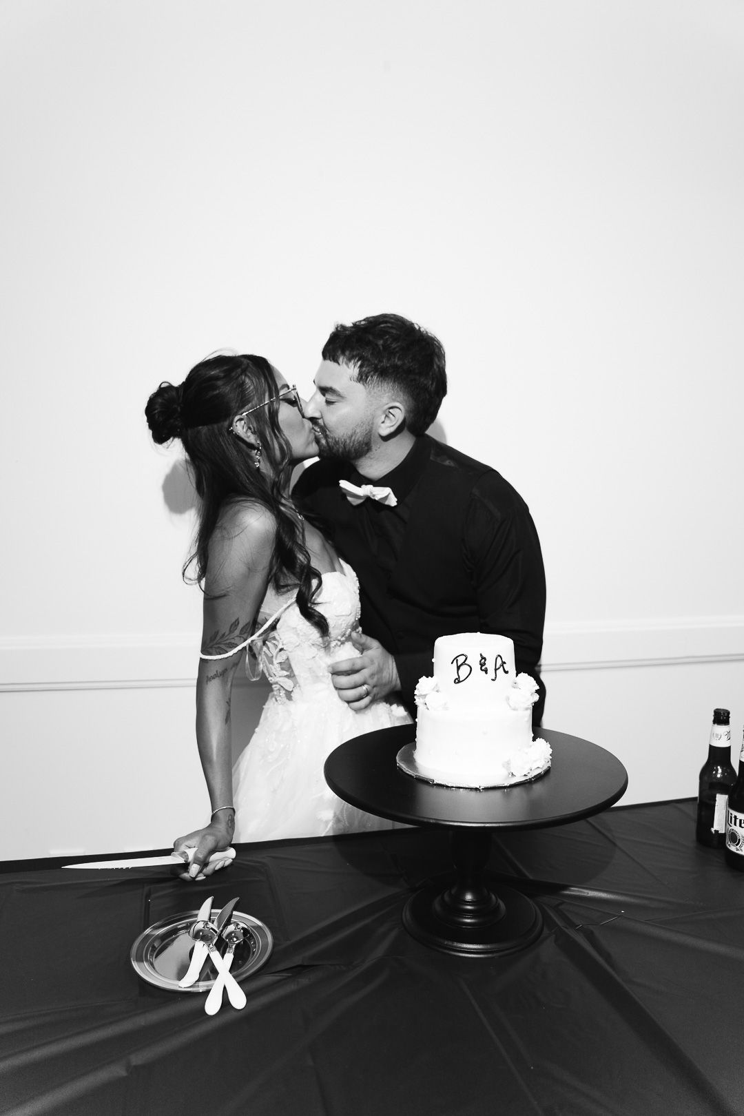Black and white photo of bride and groom kissing beside wedding cake at reception.