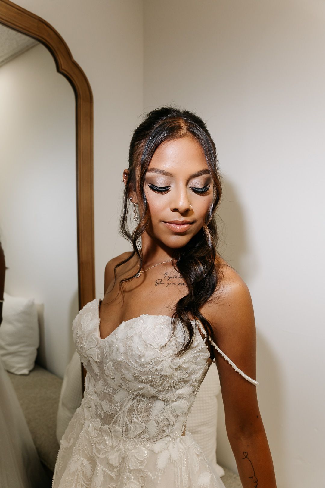 Bride in strapless lace wedding gown with elegant makeup and side-swept hair near a mirror.