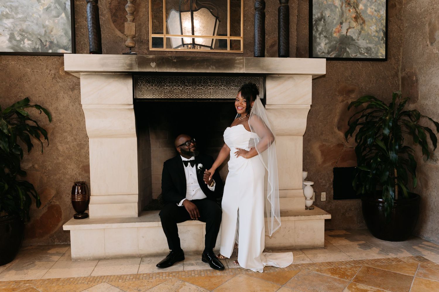 Bride in white gown and groom in black tuxedo pose elegantly beside stone fireplace in ornate venue.