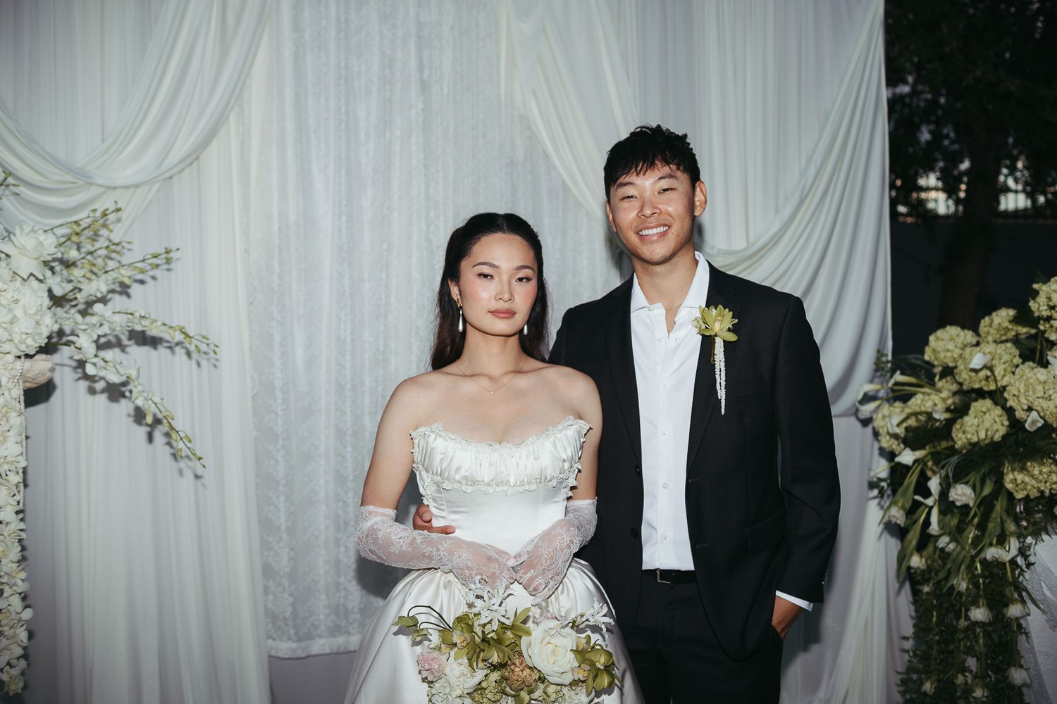 Smiling bride and groom stand together in front of a white draped backdrop with flowers.