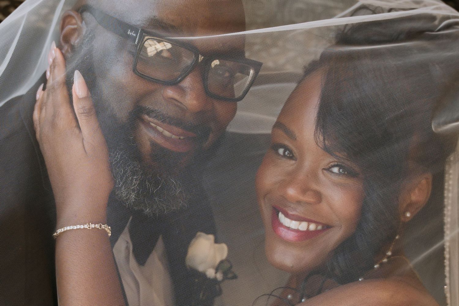 A couple smiles together under a sheer veil, the man wearing glasses and both in bridal attire.