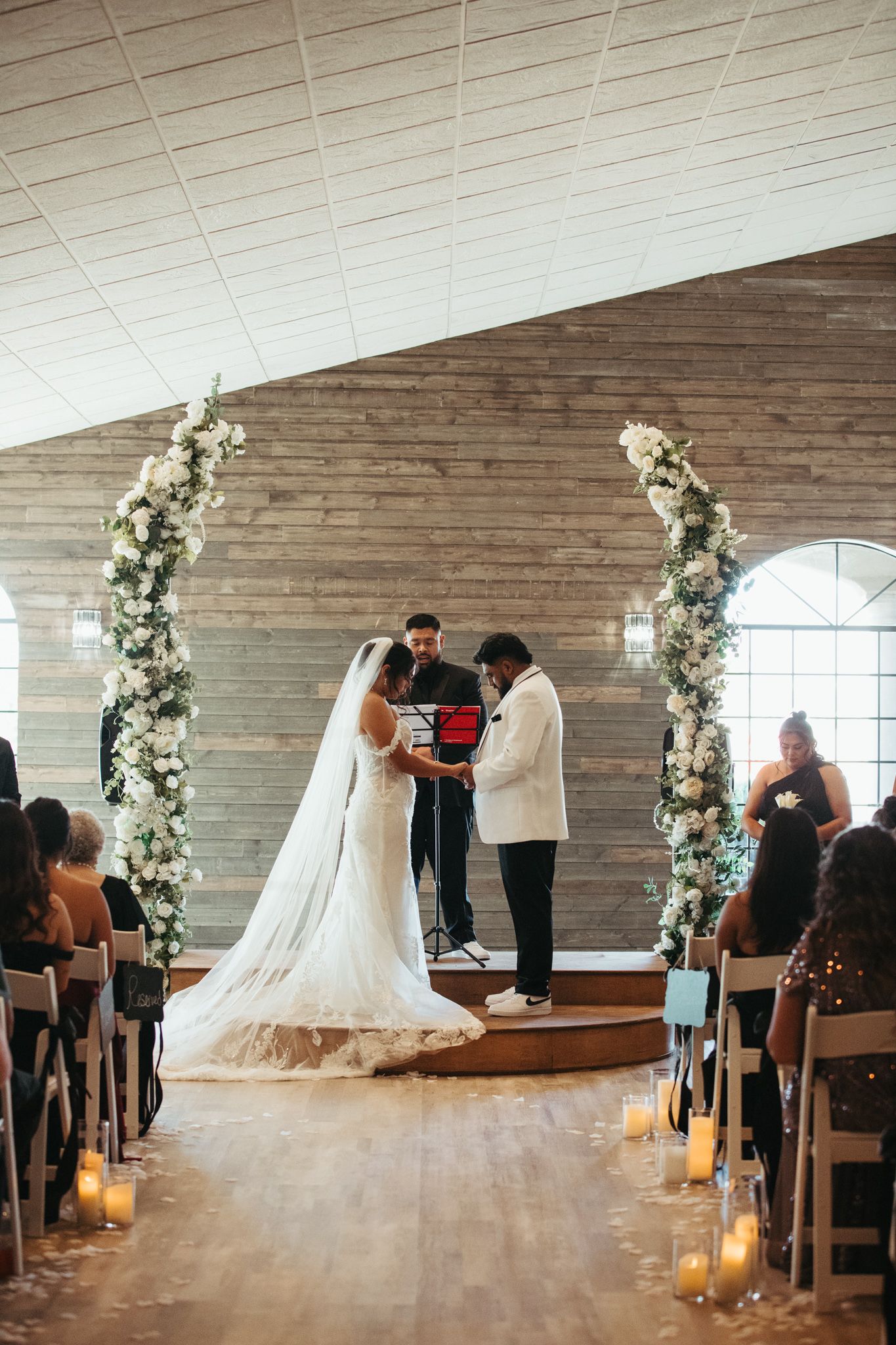 Bride and groom exchange vows at floral arch altar surrounded by white flowers and candlelit aisle.