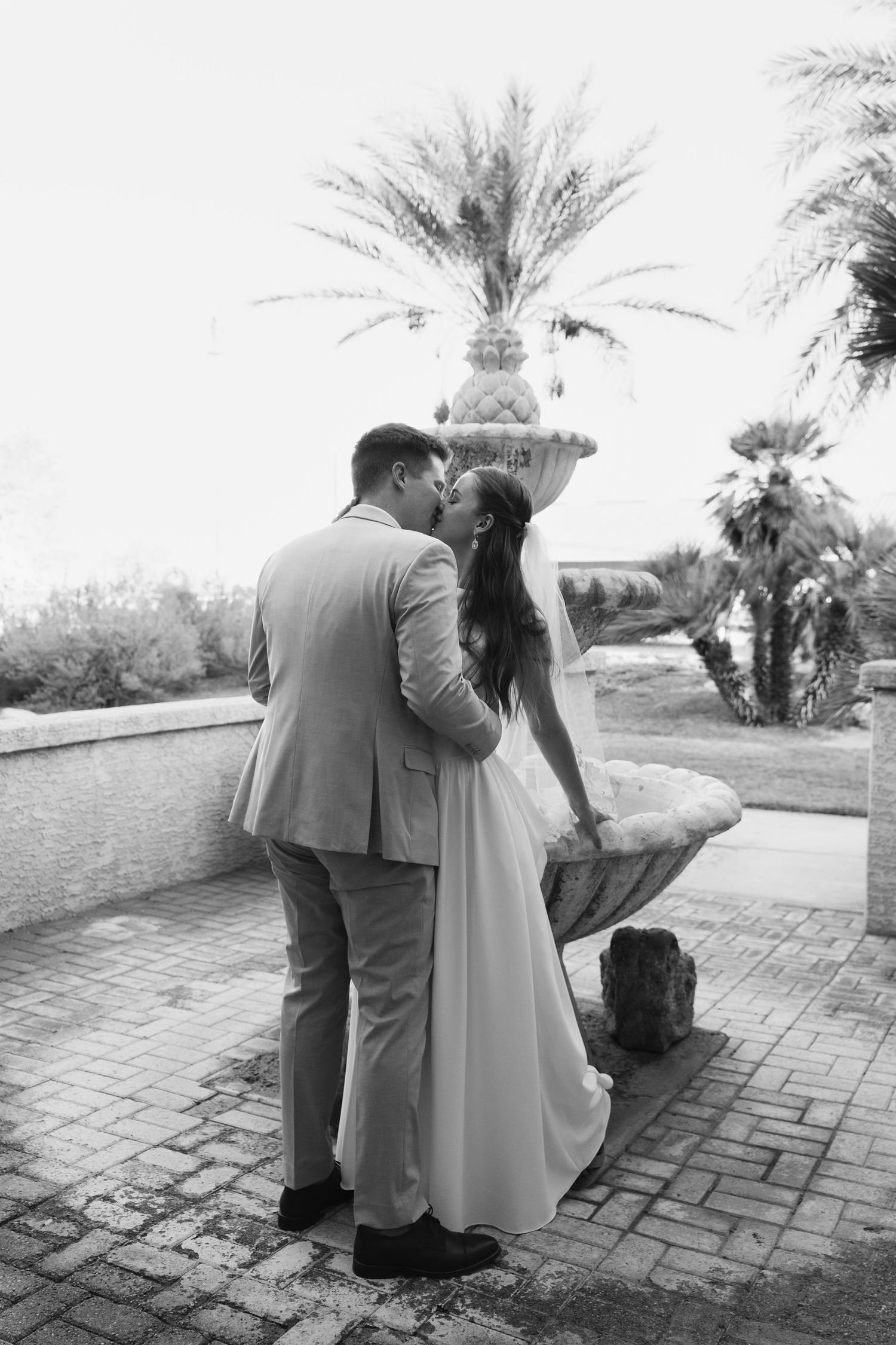 Black and white photo of bride and groom kissing near fountain with palm trees.