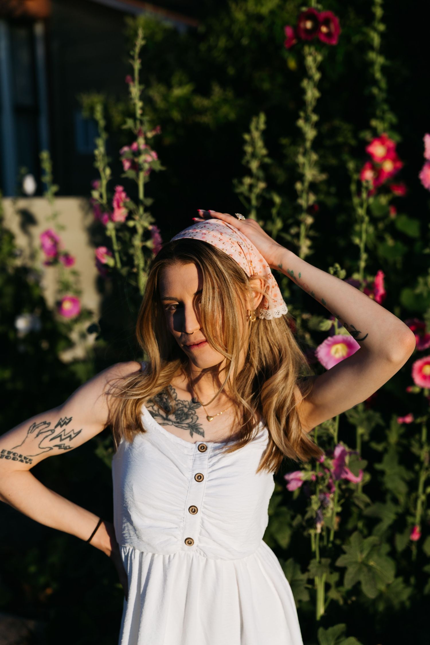 Young woman with tattoos in white sundress posing among blooming pink hollyhock flowers outdoors.