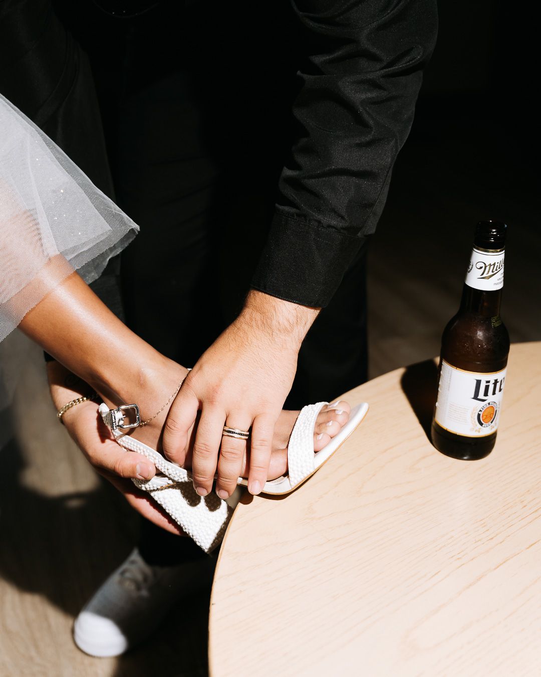 Groom cleans brides white wedding shoe on table beside a Miller Lite beer bottle at reception.