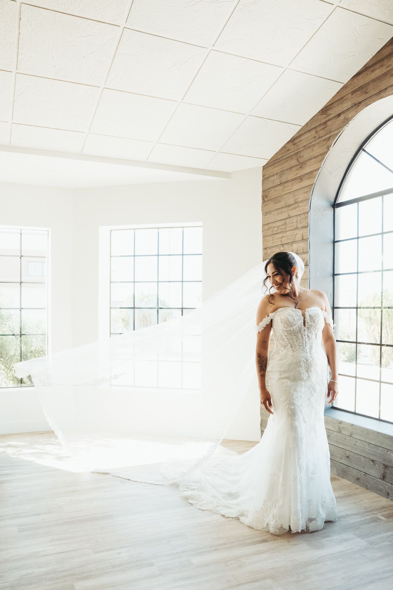Bride in off-shoulder lace mermaid gown with flowing veil poses in bright sunlit white wedding venue.