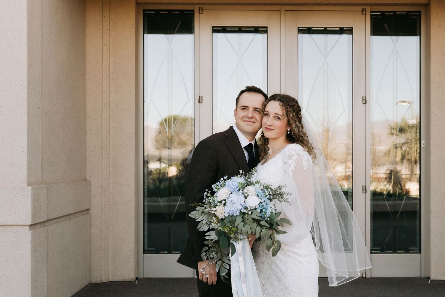Smiling bride and groom pose outside wedding venue with blue and white bouquet.