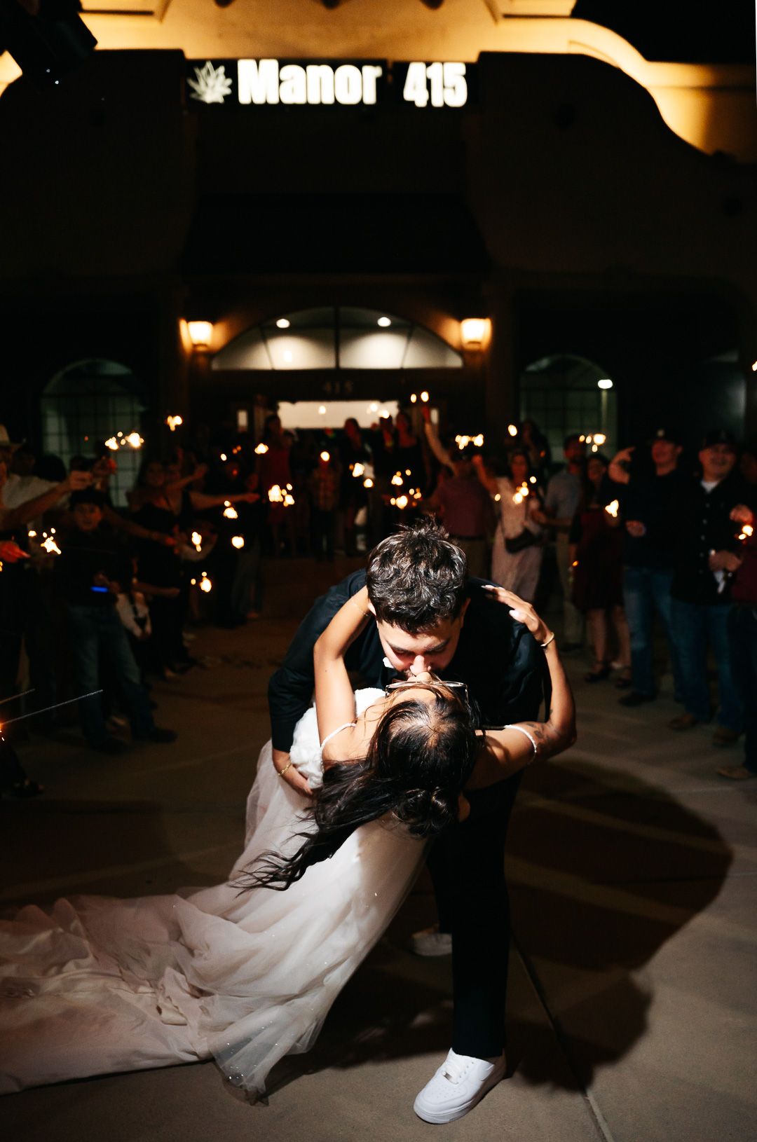 Bride and groom share a romantic dip kiss outside Manor 415 venue surrounded by guests holding sparklers at night.