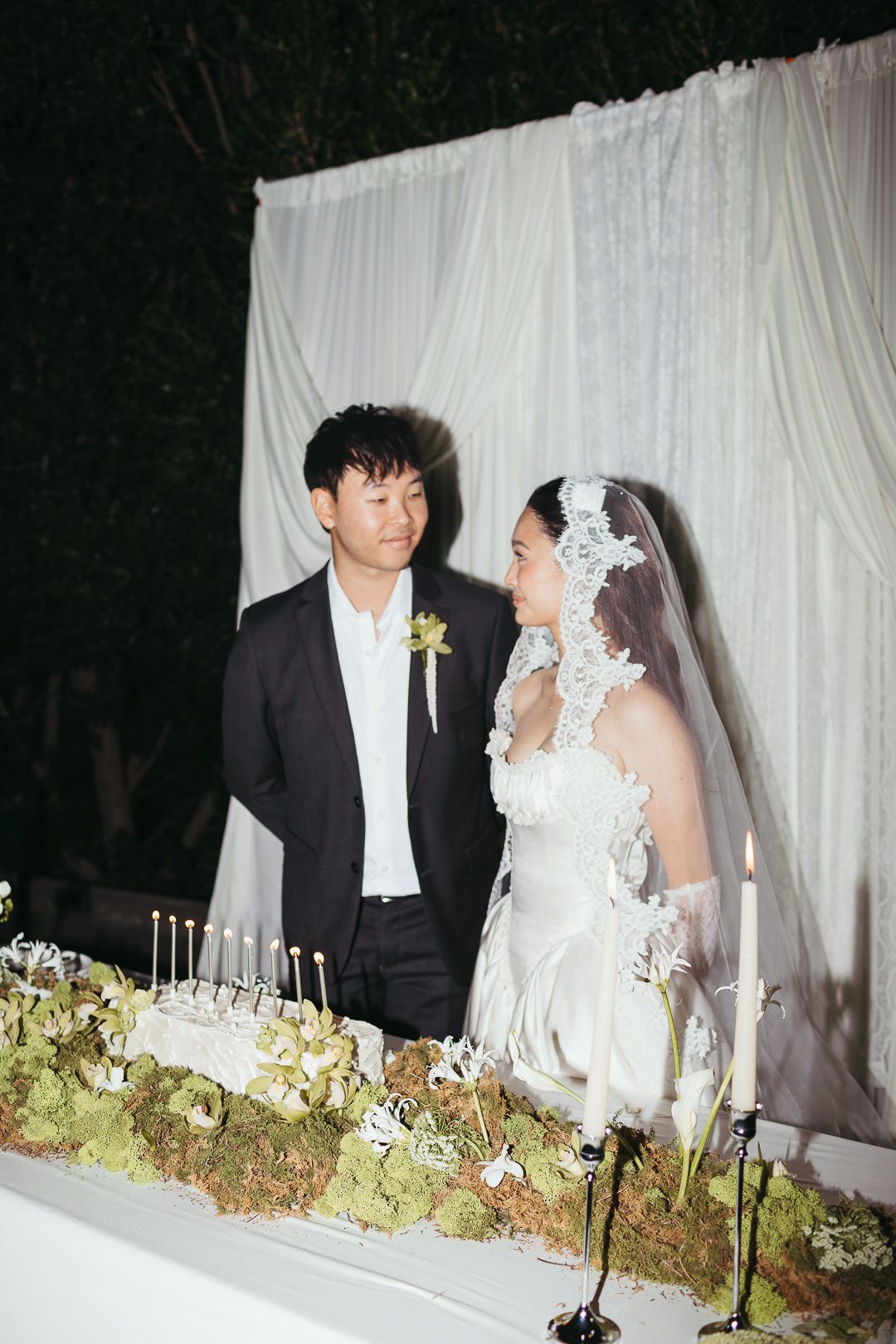 Bride in lace veil and groom exchange glances at candlelit wedding cake table before white draped backdrop.