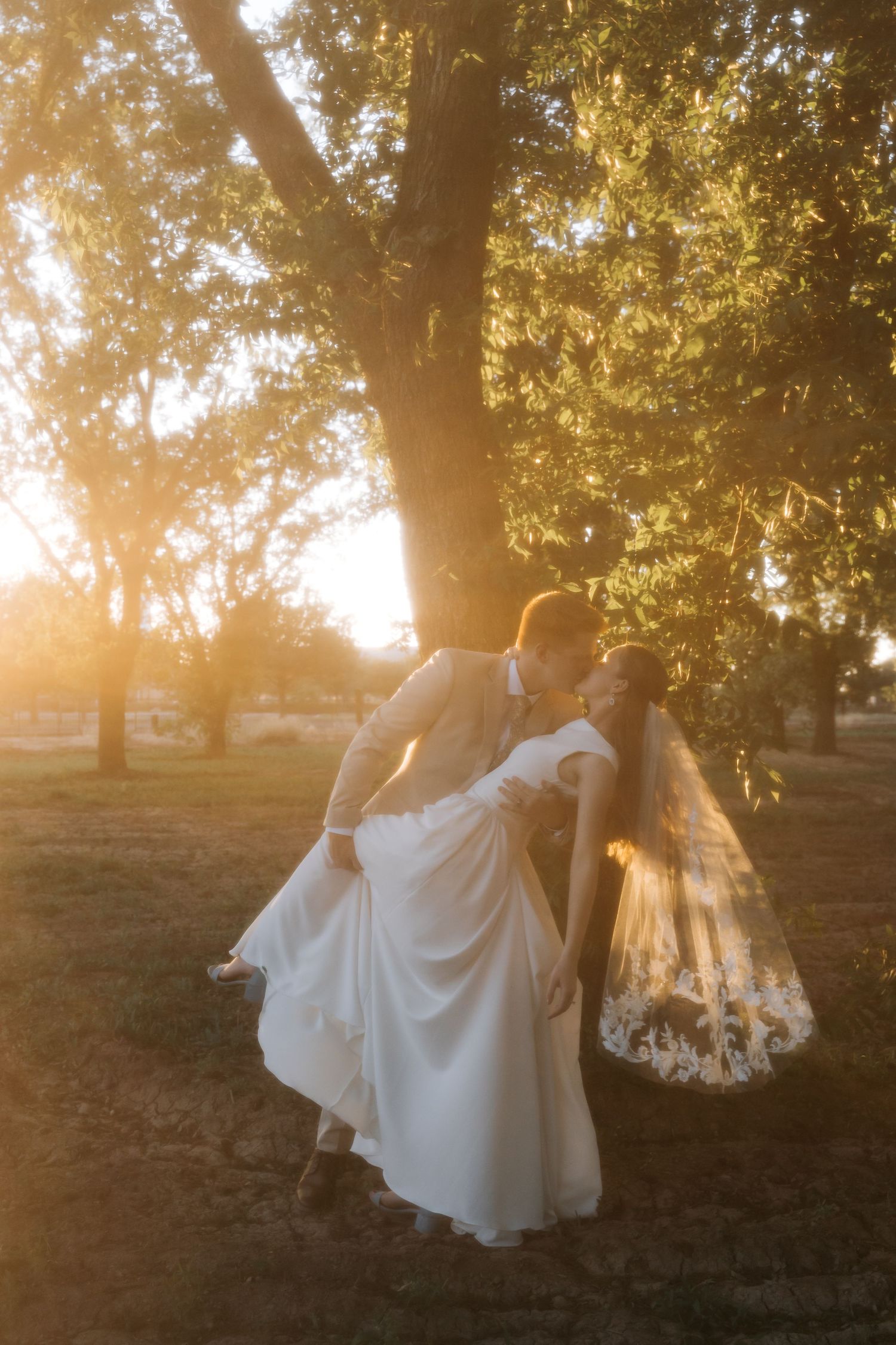 Groom lifts bride in a joyful embrace under golden sunlit trees during an outdoor wedding shoot.