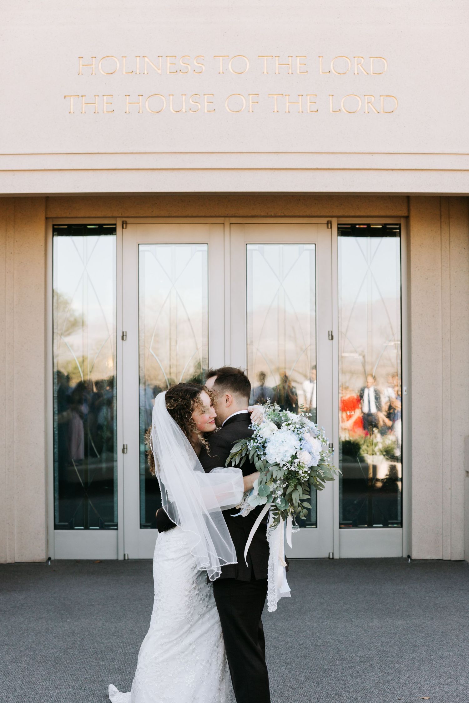 Bride and groom embrace outside LDS temple doors holding white floral bouquet.