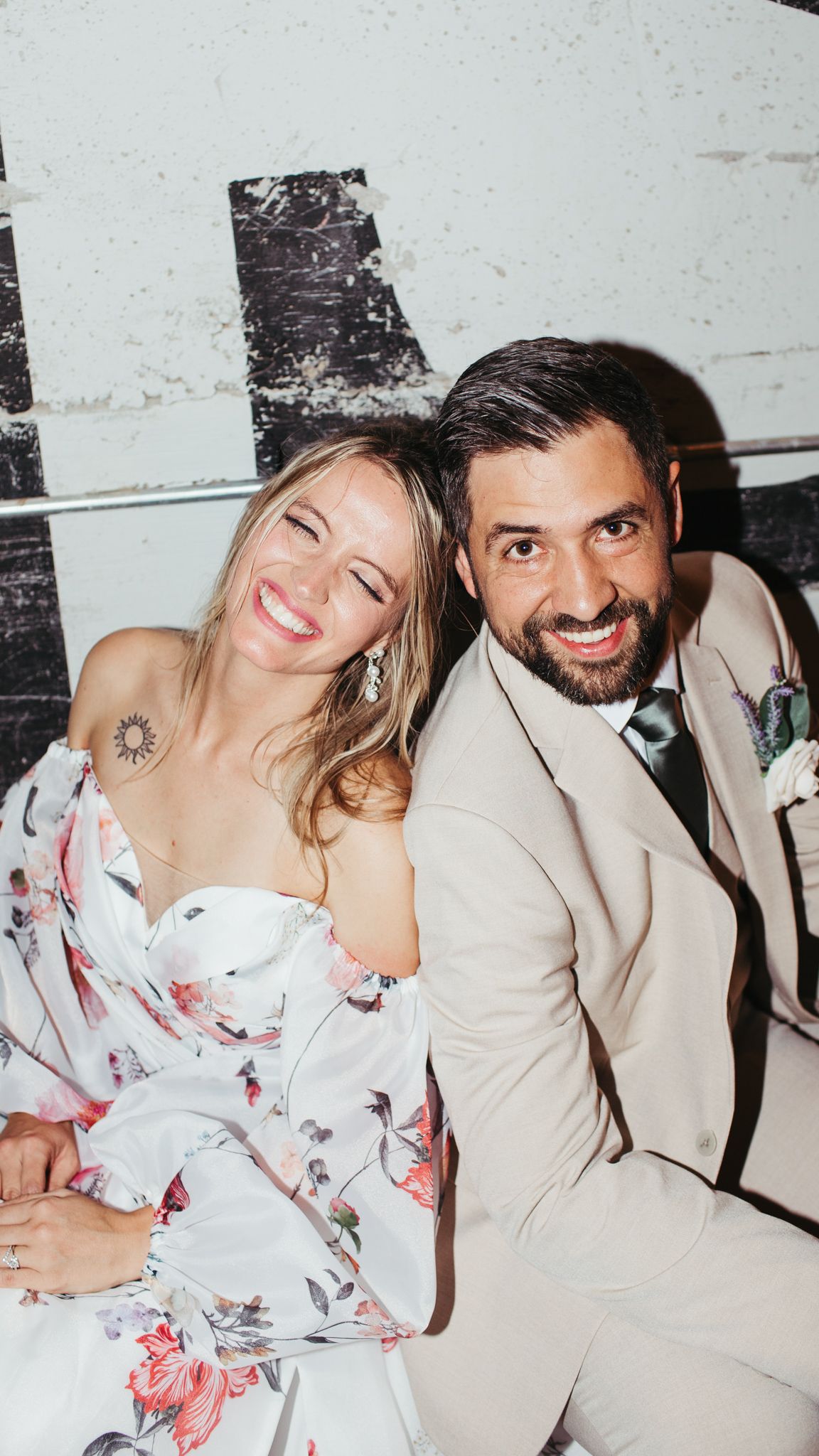 Smiling bride in floral dress and groom in beige suit sitting together at wedding reception.