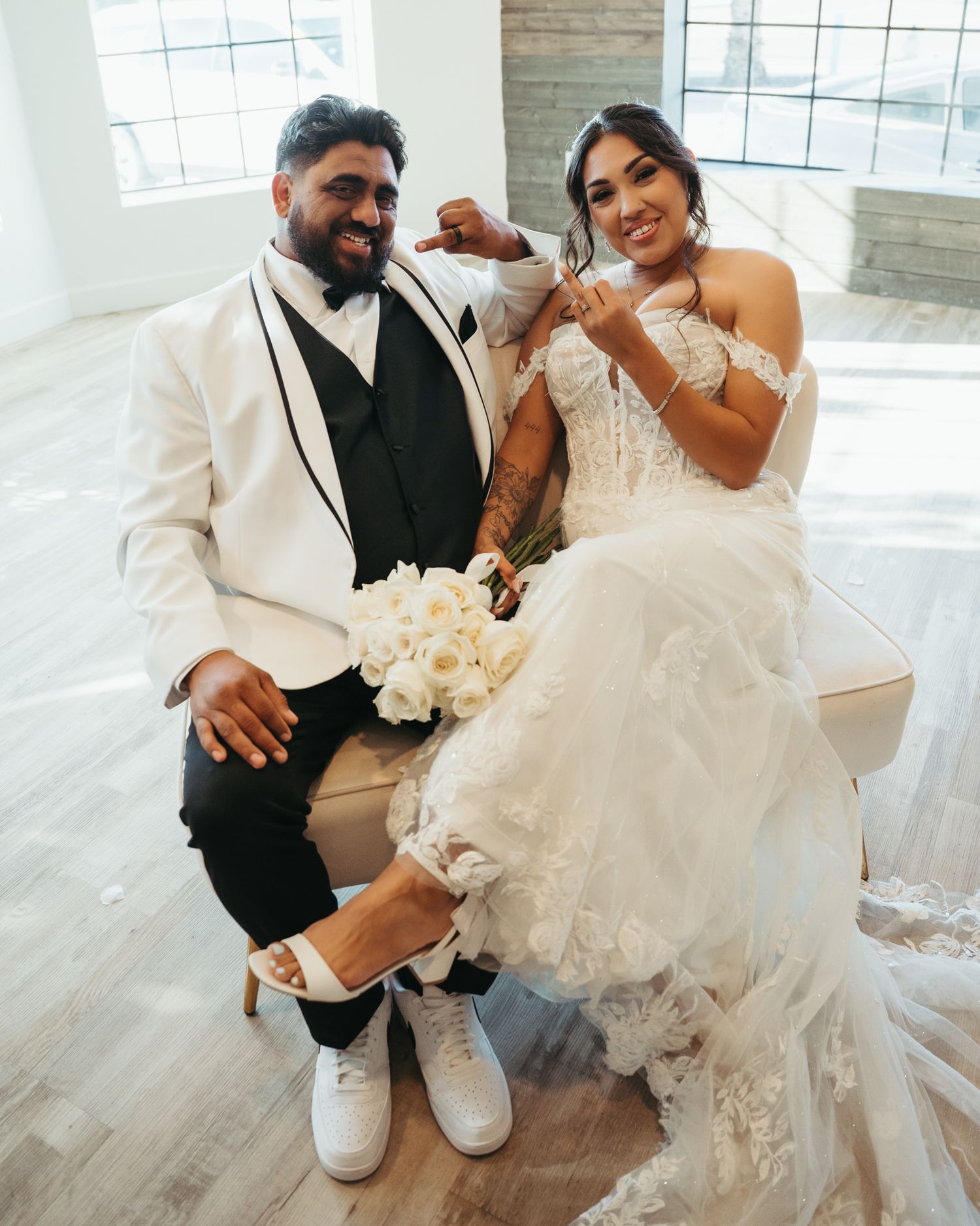 Bride and groom sitting together smiling, groom in white tuxedo and bride in lace wedding dress holding white bouquet.