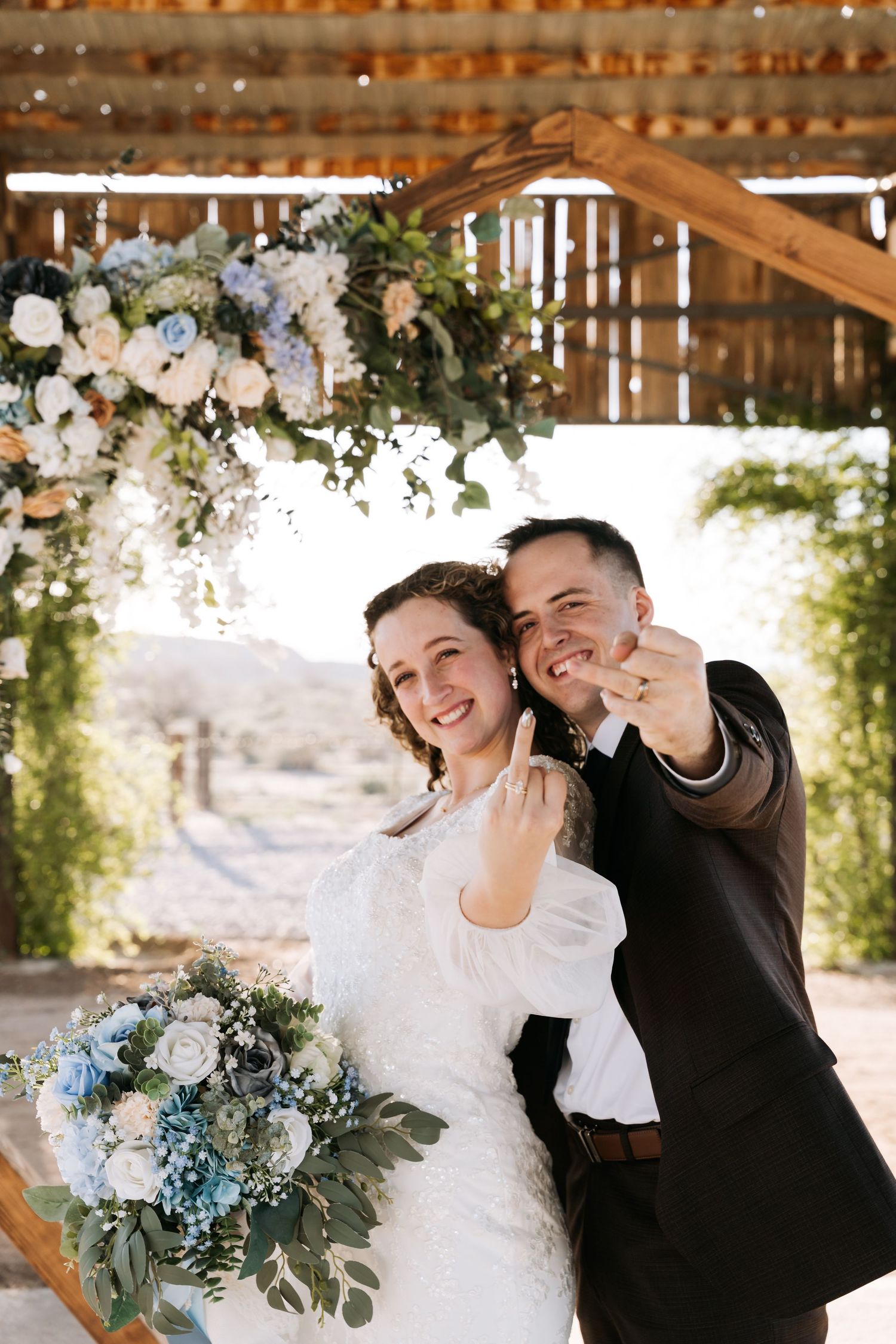 Smiling newlyweds show off wedding rings under floral arch with blue and white flowers at outdoor ceremony.