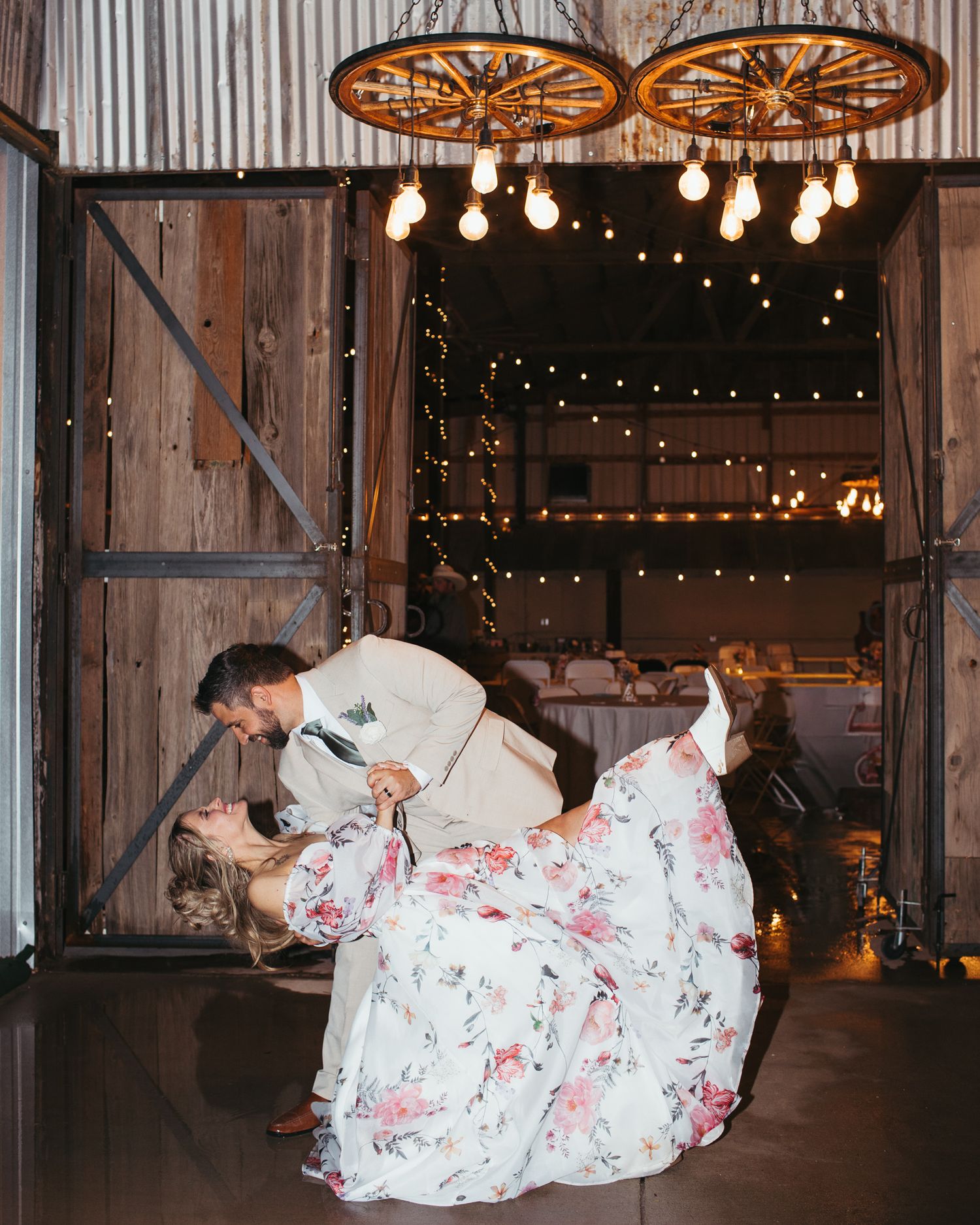Groom dips bride in floral wedding dress during first dance in a rustic barn with string lights.
