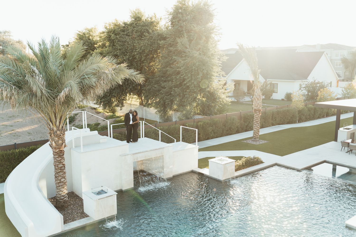Aerial view of couple standing on white bridge overlooking resort pool surrounded by palm trees at sunset.