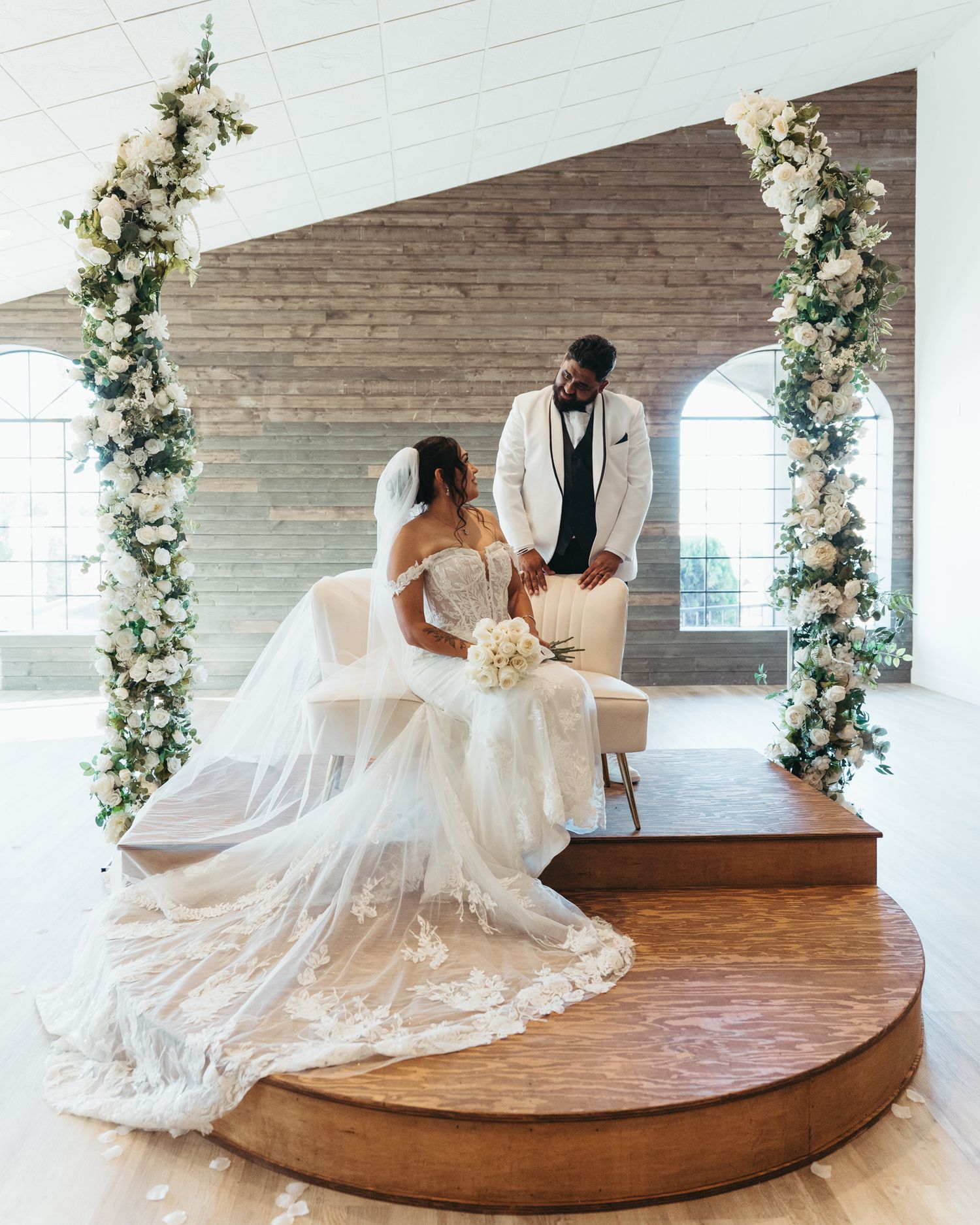 Bride in lace gown and groom in white suit share intimate moment on wooden altar with floral columns.