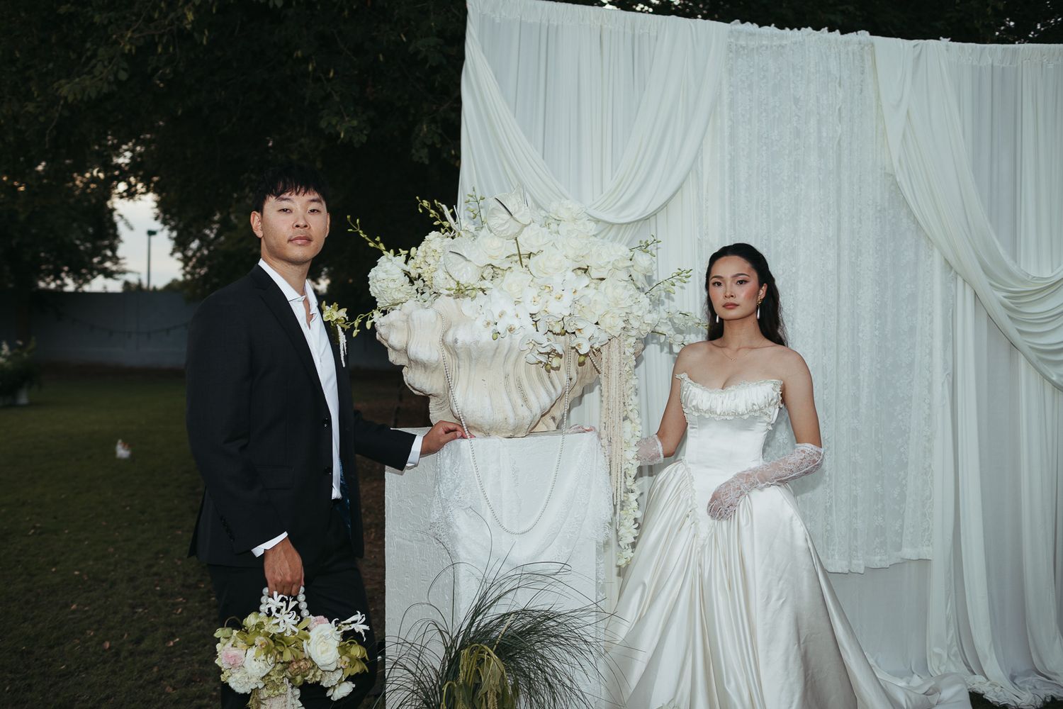 Bride and groom pose elegantly before a white draped backdrop with white floral arrangements.