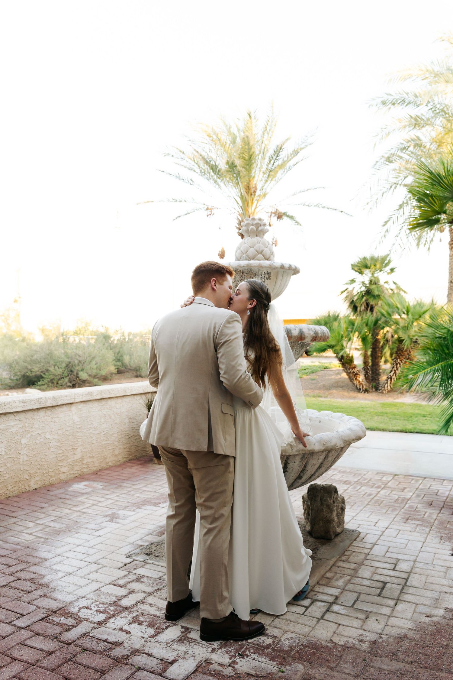 Bride and groom share a romantic kiss near a stone fountain surrounded by palm trees at sunset.