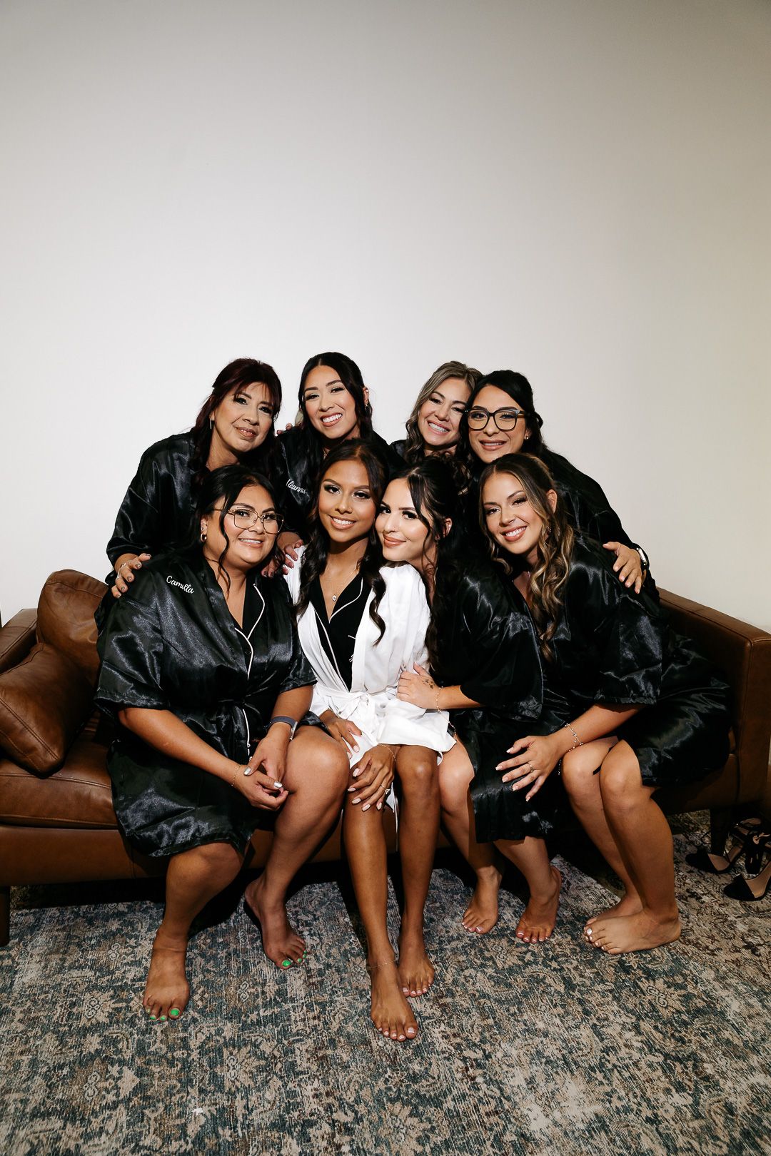 Bride in white robe surrounded by seven bridesmaids in black satin robes on a leather sofa.