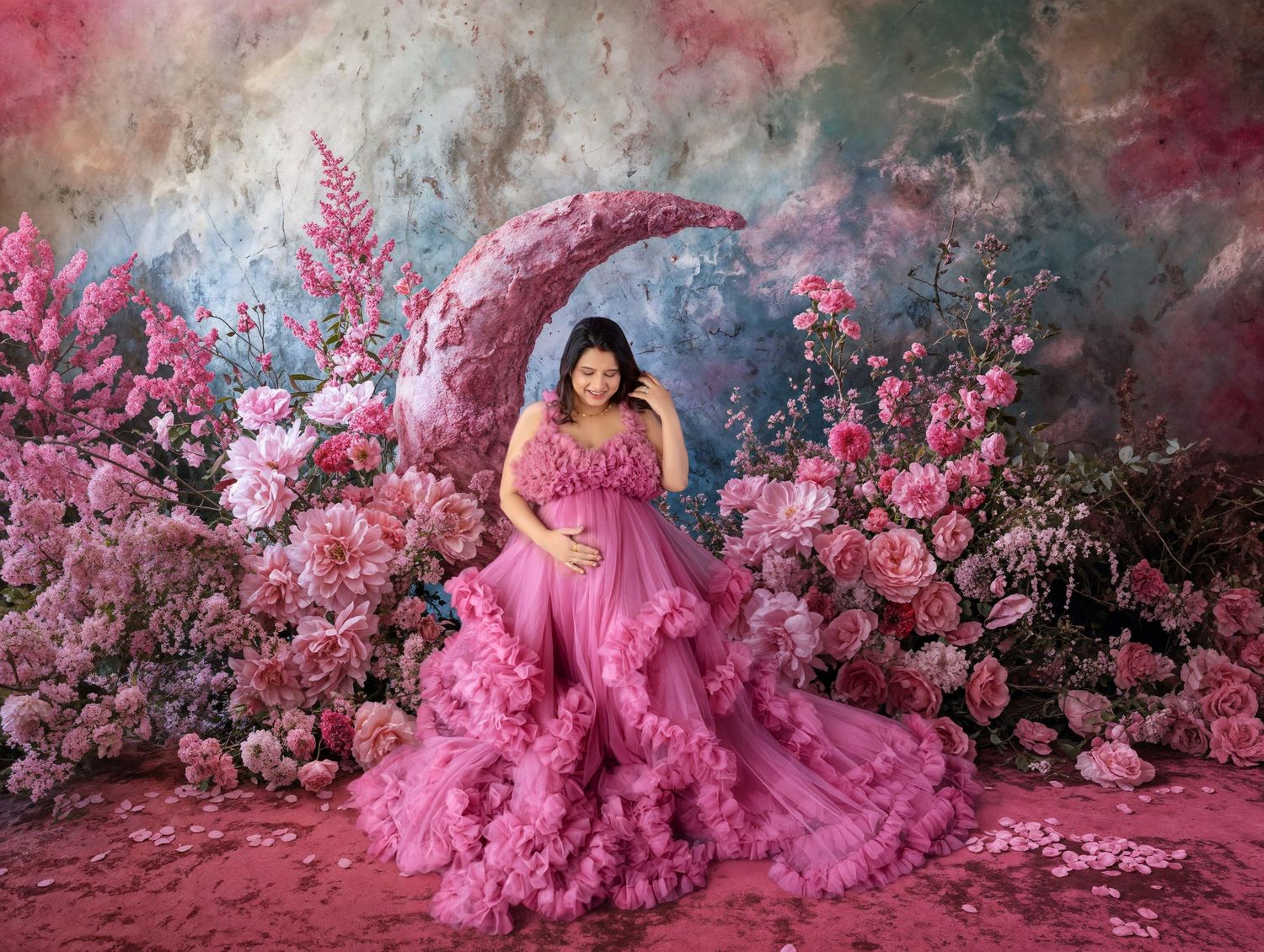 Pregnant woman in pink ruffle gown poses on floral moon prop surrounded by pink flowers in dreamy maternity photo.